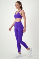 A woman is standing and smiling, wearing matching purple activewear—a sports bra and DRIVEN Gym Apparel Classic Full Length Tights (sapphire). She also has on white sneakers. Her hair is tied back in a ponytail as she poses against a plain white background, showcasing her chic gym wear.