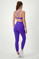 A woman with a ponytail is standing and facing away from the camera, dressed in a matching set of purple fitness apparel with crisscross straps and DRIVEN Gym Apparel's Classic Full Length Tights (sapphire). She is also wearing white sneakers.