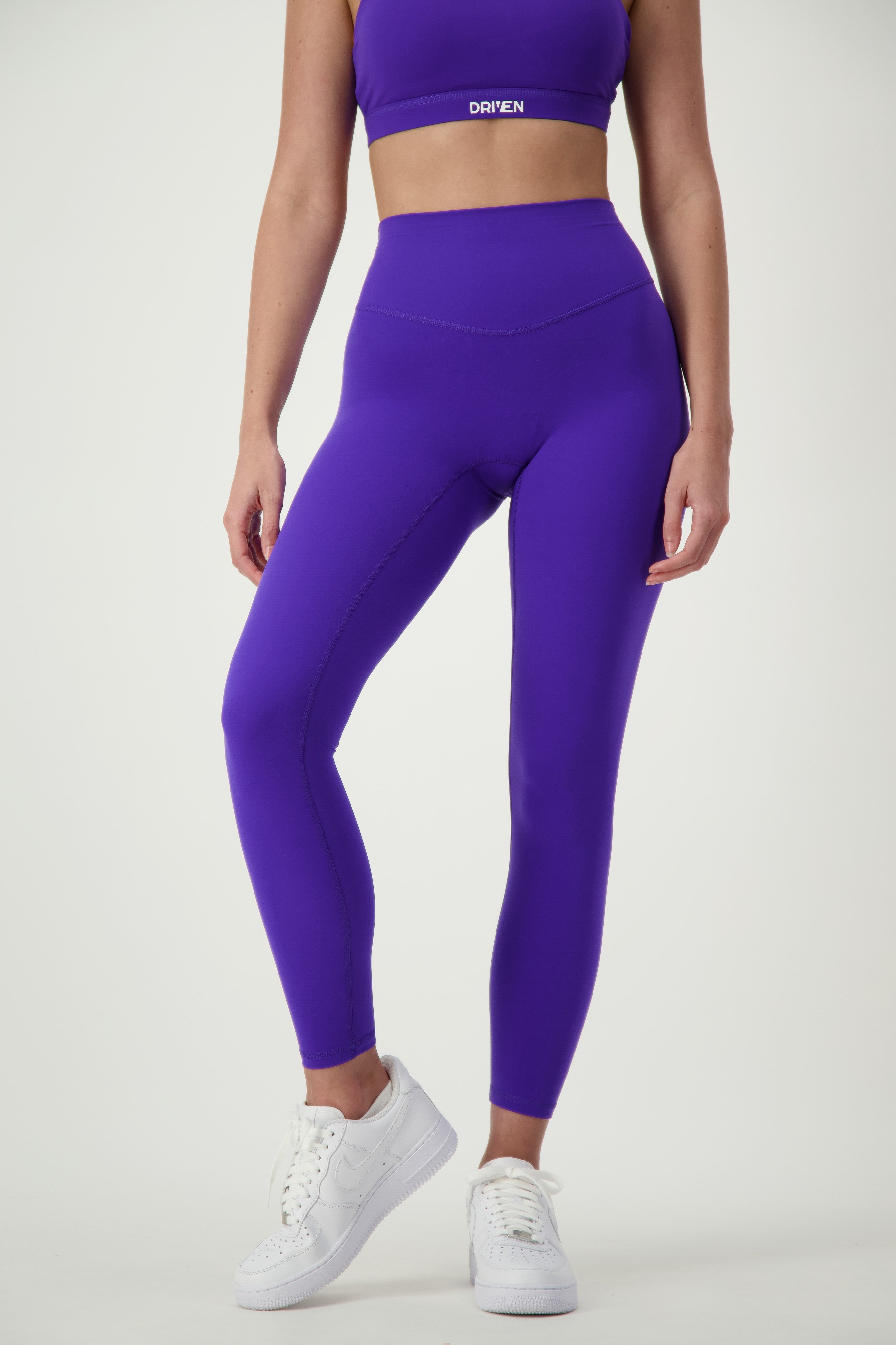 A person is shown from the shoulders down, dressed in vibrant purple athleisure, including a sports bra and matching high-waisted Classic Full Length Tights (sapphire). The outfit is complemented by white sneakers and set against a plain white background. The sports bra proudly displays the word "DRIVEN Gym Apparel" on the chest.