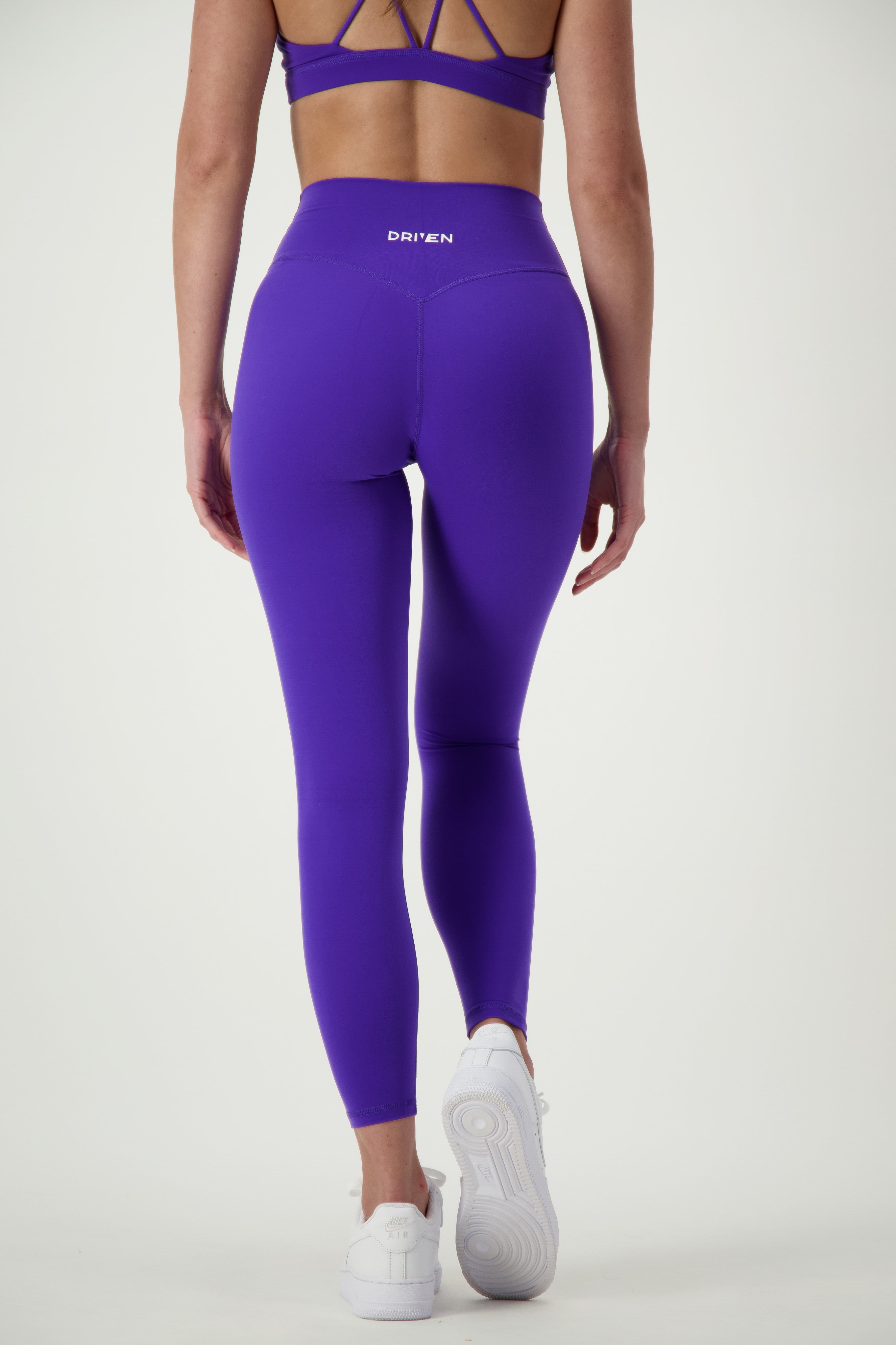 A person in a purple athletic outfit, seen from the back, is standing on a neutral background. They are wearing a sports bra and Classic Full Length Tights (sapphire) with the brand name "DRIVEN Gym Apparel" visible on the waistband. This stylish activewear ensemble is completed with white sneakers.