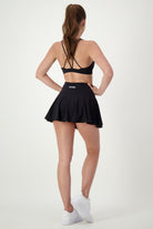 A woman with long brown hair in a ponytail is standing with her back to the camera. She is wearing gym wear: a black sports bra with criss-cross straps, a black Classic Mini Skort from DRIVEN Gym Apparel, and white sneakers. Her hands are on her hips as she stands against a plain white background.