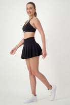 A person with a ponytail is posing in athleisure, featuring a black sports bra, DRIVEN Gym Apparel's black Classic Mini Skort, and white sneakers. They are looking over their shoulder and smiling, standing in a light setting with a plain background.