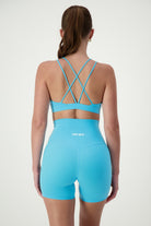 A person with long brown hair in a ponytail is facing away from the camera, wearing matching light blue activewear: a sports bra with crisscross straps and high-waisted Classic Midi Shorts (sky). The word "DRIVEN Gym Apparel" is printed on the back of the shorts. The background is white.