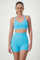 A smiling woman with long hair in a ponytail wears matching light blue fitness apparel consisting of a sports bra and Classic Midi Shorts (sky). The sports bra, adorned with the text "DRIVEN," highlights her commitment to activewear. The background is plain white.
