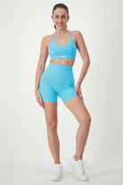 A woman stands on a white background, smiling while wearing light blue fitness apparel consisting of a sports bra and Classic Midi Shorts (sky) by DRIVEN Gym Apparel. Her hair is pulled back, and she completes the look with white sneakers. The matching outfit features a logo on the sports bra.
