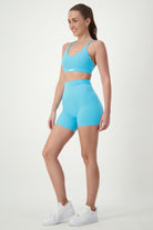 A woman stands against a plain white background, wearing stylish activewear: a matching light blue sports bra and high-waisted **Classic Midi Shorts (sky)** from **DRIVEN Gym Apparel**. She has her hair tied back and is smiling, completing the look with white sneakers.