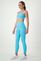 A woman with long hair tied back stands facing slightly to the side, smiling. She is wearing matching light blue fitness apparel consisting of a sports bra and DRIVEN Gym Apparel's Classic Full Length Tights (sky), along with white sneakers. The background is plain white.