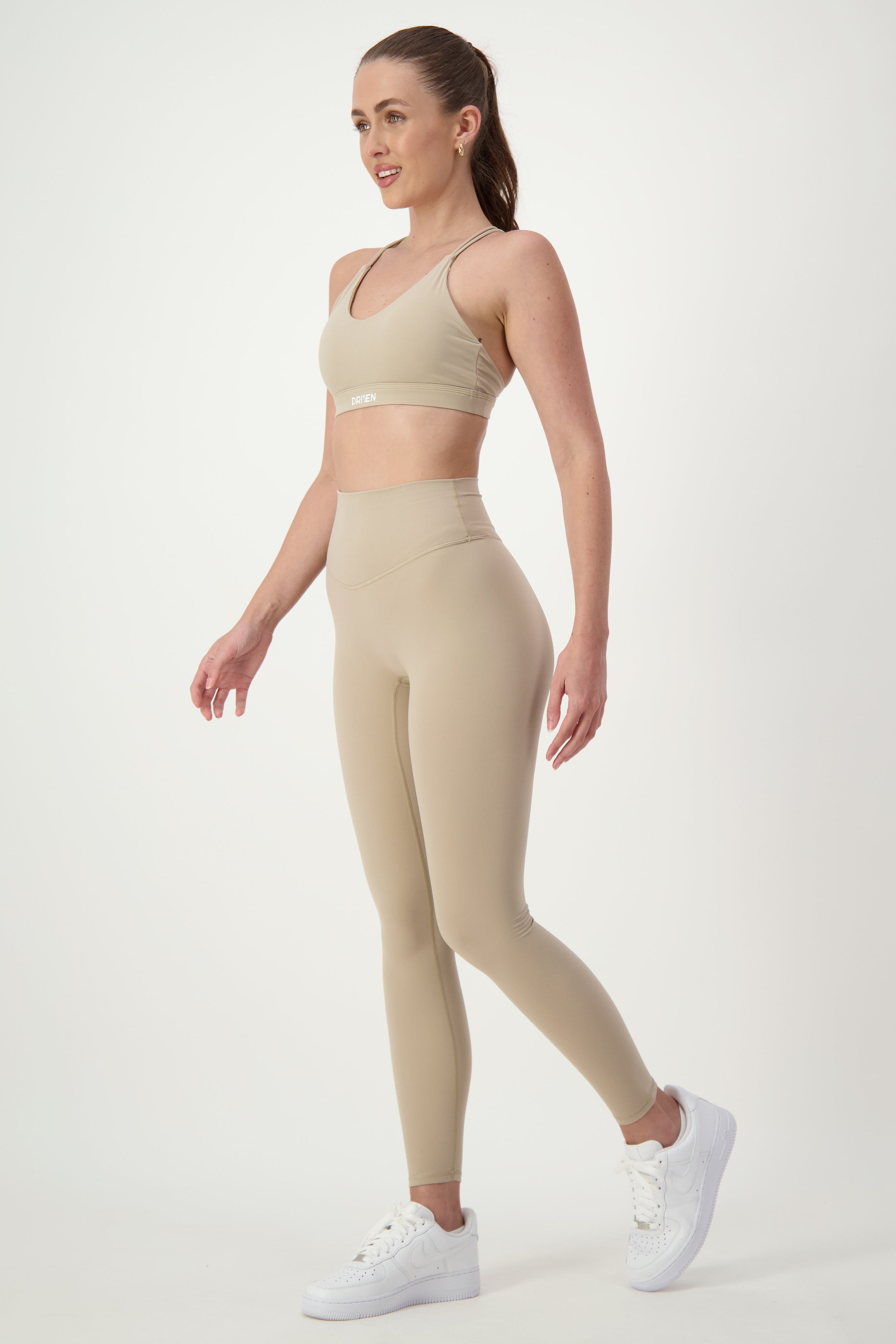A woman stands against a plain white background, wearing matching beige gym wear consisting of a sports bra and DRIVEN Gym Apparel's Classic Full Length Tights (taupe). She also wears white sneakers. Her hair is tied back, and she is posed in a casual stance with one leg slightly lifted.