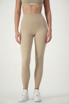 A person is wearing a matching beige sports bra and Classic Full Length Tights (taupe) set, paired with white sneakers. The word "DRIVEN" is visible on the band of the sports bra, highlighting its premium fitness apparel. The background is a plain, white studio backdrop.