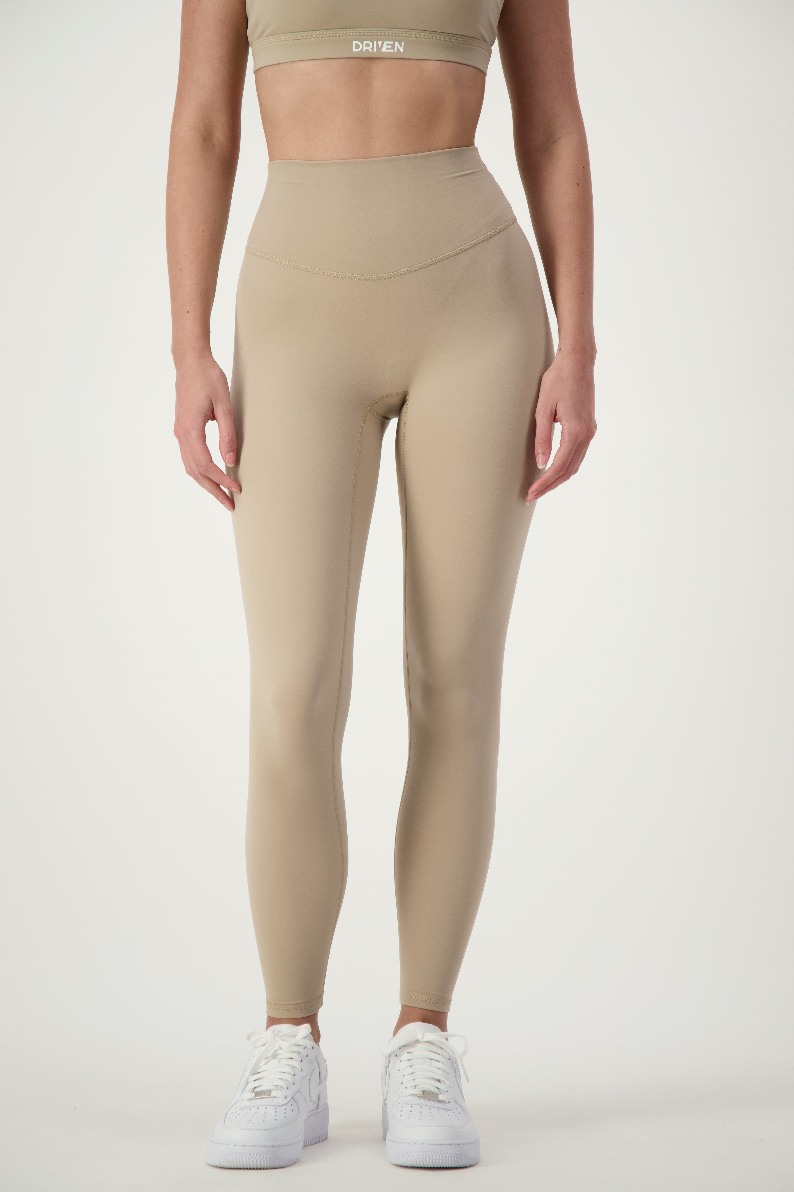 A person is wearing a matching beige sports bra and Classic Full Length Tights (taupe) set, paired with white sneakers. The word "DRIVEN" is visible on the band of the sports bra, highlighting its premium fitness apparel. The background is a plain, white studio backdrop.