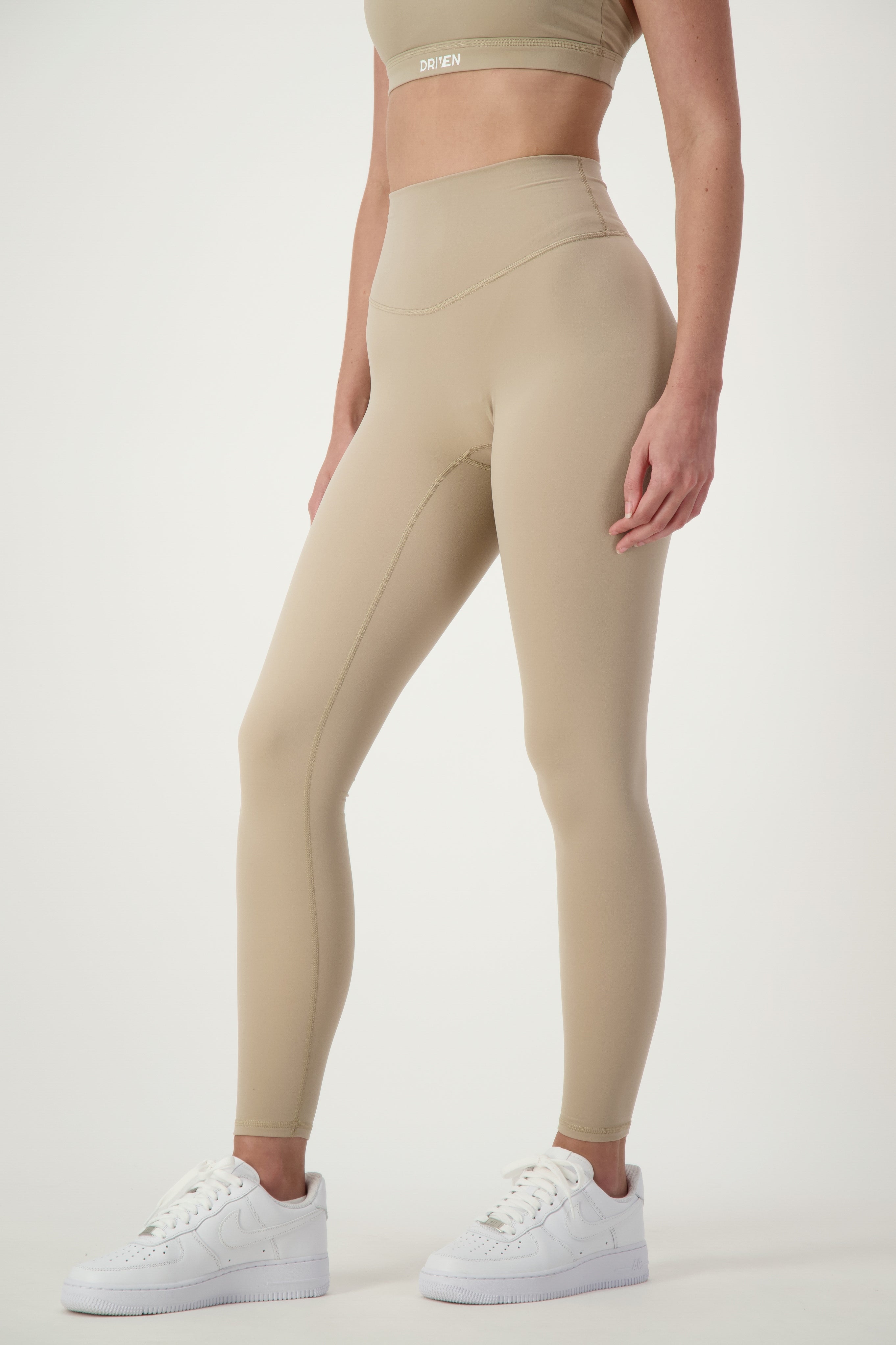 A person wearing DRIVEN Gym Apparel's taupe Classic Full Length Tights and a matching sports bra, epitomizing athleisure. They are also sporting white athletic shoes against a plain, light-colored background. The person's head is not visible in the image.