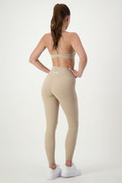 A woman with long brown hair in a ponytail is standing against a plain white background. She is wearing beige fitness apparel, which includes a sports bra, matching Classic Full Length Tights (taupe) by DRIVEN Gym Apparel with "GIVVISEL" written on the back, and white sneakers. She is looking over her left shoulder.