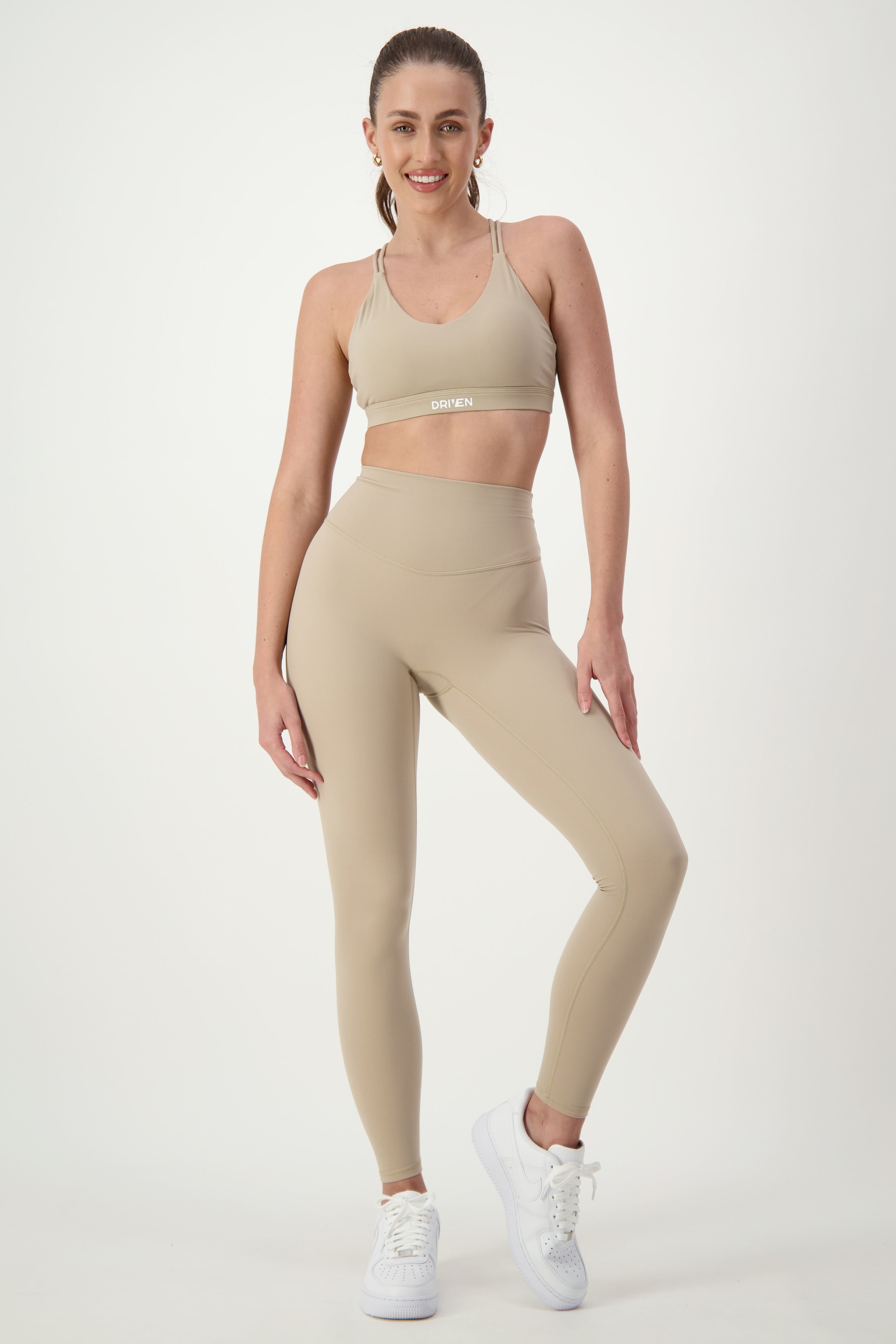 A person with pulled-back hair is posing against a white background, showcasing trendy fitness apparel in a beige sports bra and matching high-waisted Classic Full Length Tights (taupe) from DRIVEN Gym Apparel. They are wearing white sneakers, smiling confidently, with one hand on their hip and the other resting by their side.