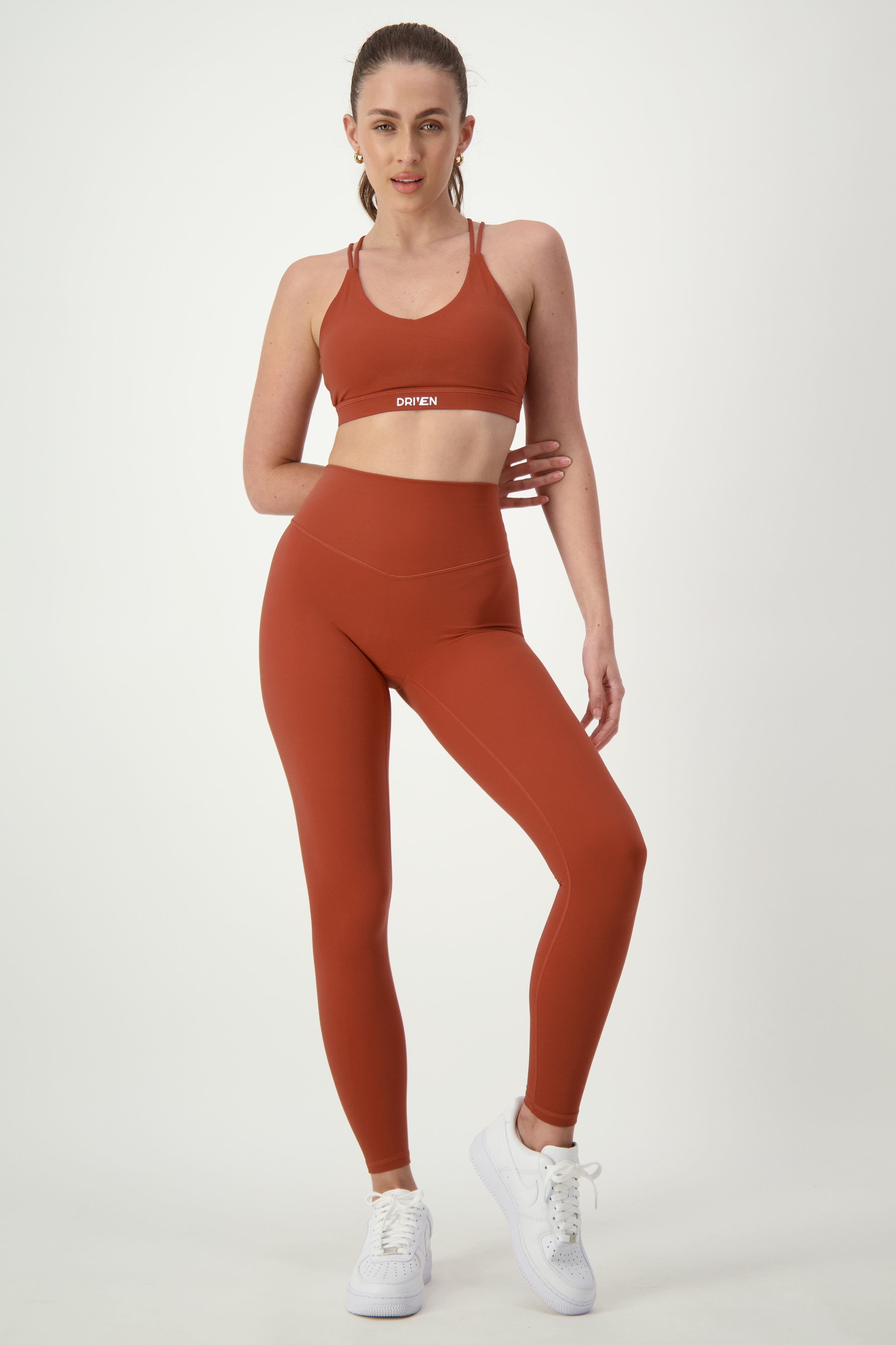 A woman wearing burnt orange activewear, consisting of a sports bra and matching DRIVEN Gym Apparel Classic Full Length Tights (umber), stands against a plain white background. She has her hair pulled back, is wearing white sneakers, and one hand is resting on her thigh while the other is on her hip.