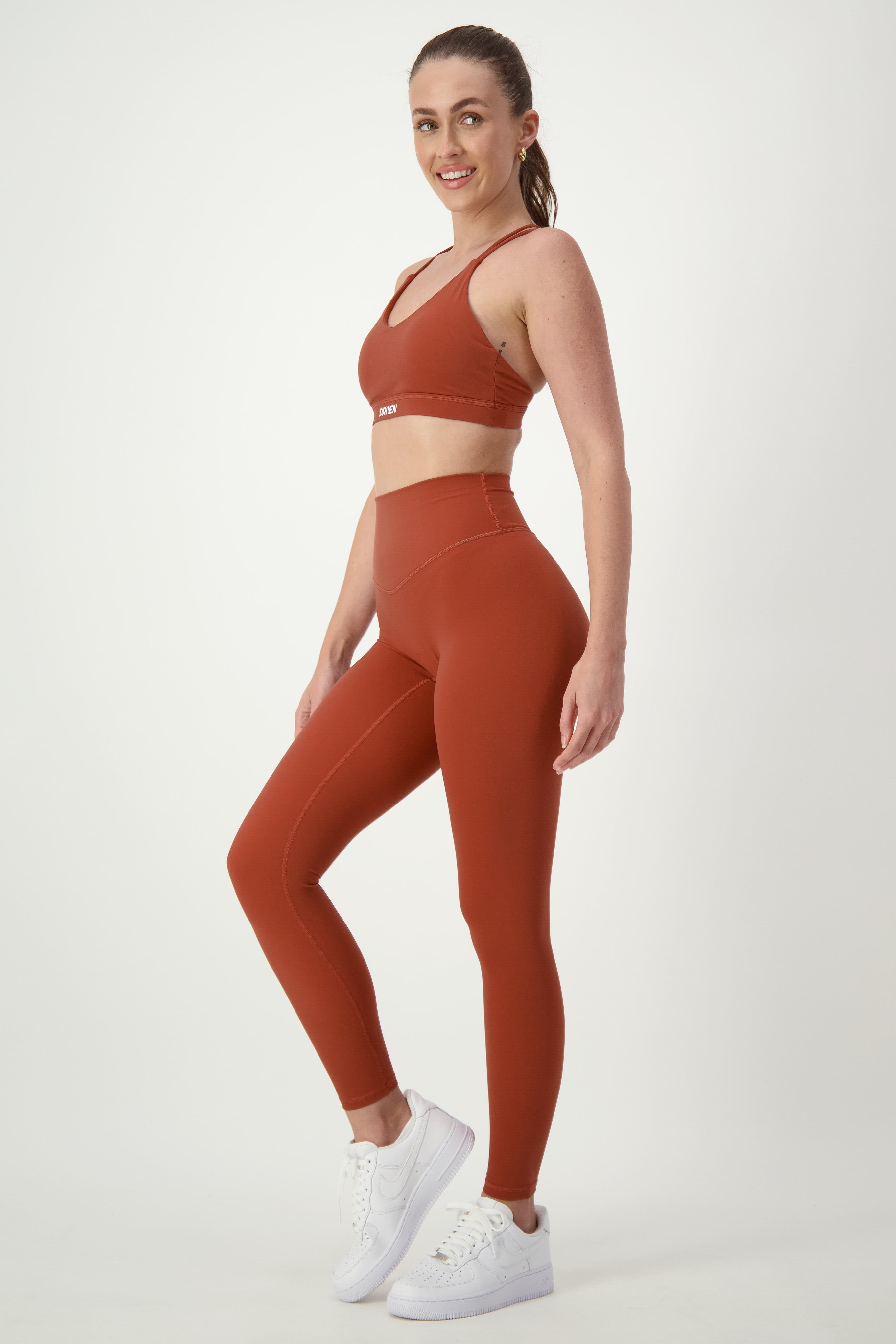 A woman stands against a plain background, modeling DRIVEN Gym Apparel's matching set of rust-colored gym wear, including a sports bra and Classic Full Length Tights (umber). She is wearing white sneakers and smiling at the camera, with her hair tied back in a ponytail.