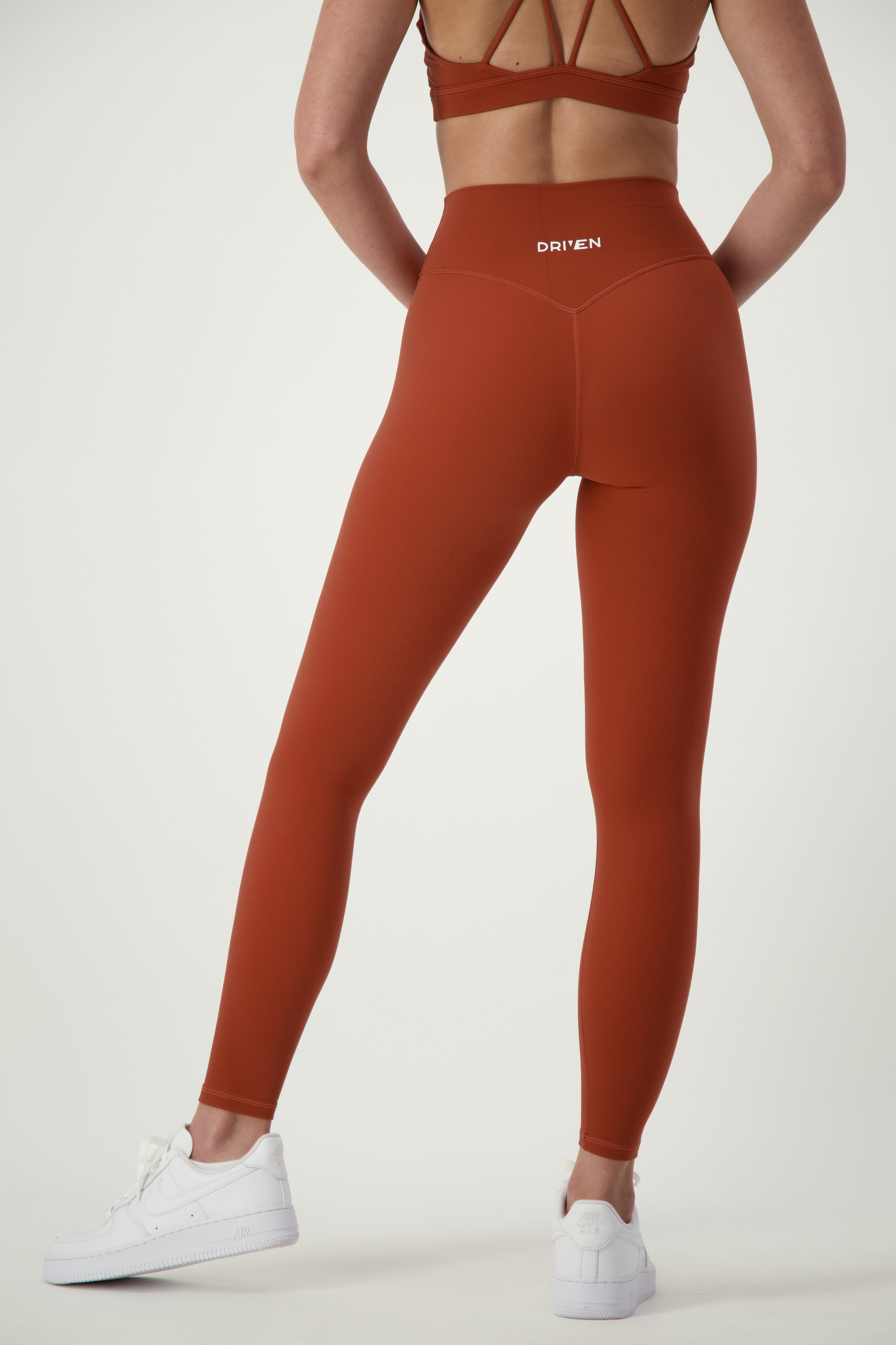 A person is shown from the back wearing burnt orange high-waisted leggings and a matching sports bra, epitomizing stylish fitness apparel. The Classic Full Length Tights (umber) by DRIVEN Gym Apparel have the word "DRIVEN" printed on the waistband. Paired with white sneakers, this activewear outfit stands out against a plain, light-colored background.