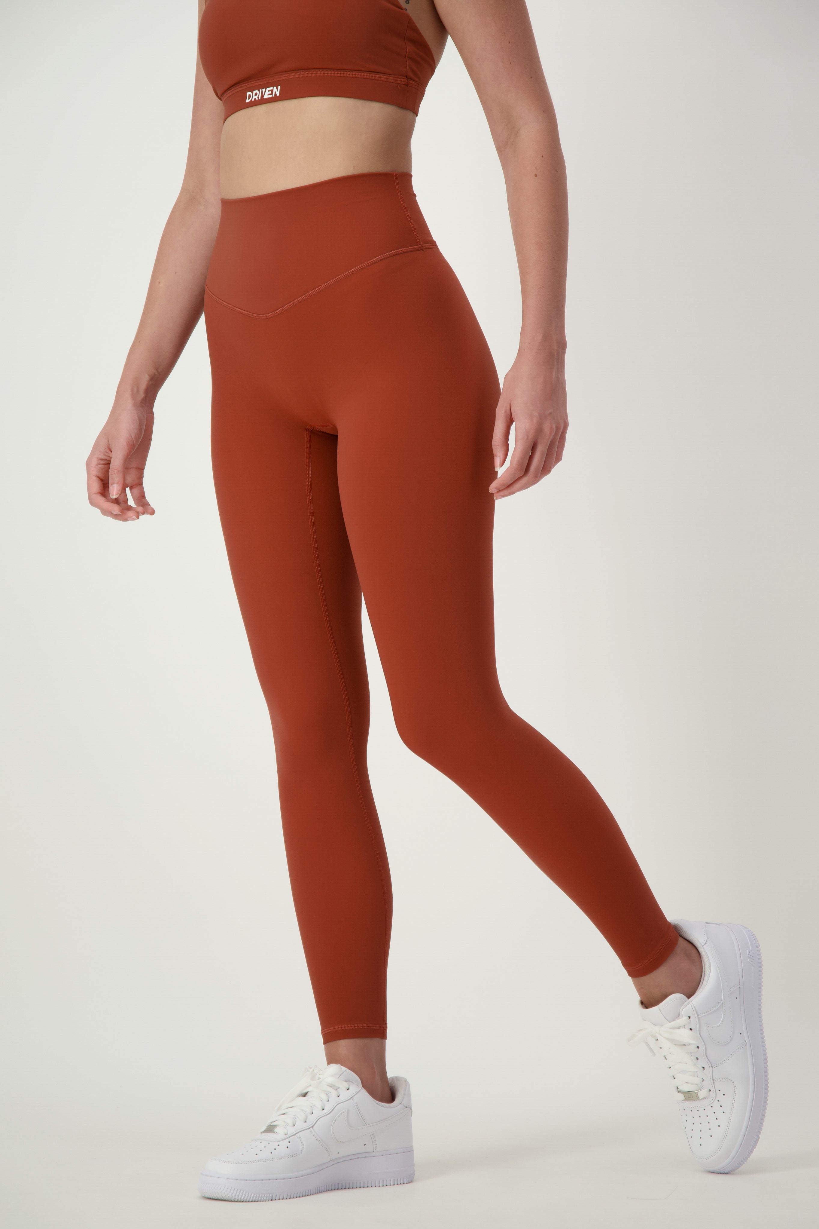 A person is showcasing athleisure in a matching burnt orange sports bra and DRIVEN Gym Apparel's Classic Full Length Tights (umber). Complemented by white sneakers, the photo is cropped from the shoulders down against a plain light background.