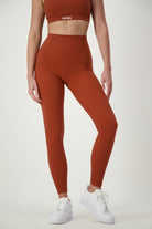 A woman stands against a plain background wearing rust-colored fitness apparel, consisting of a sports bra with the word "DRIVEN" printed on it and matching Classic Full Length Tights (umber) from DRIVEN Gym Apparel, paired with white sneakers. She is posing with her left hand resting on her hip and her right arm hanging by her side.