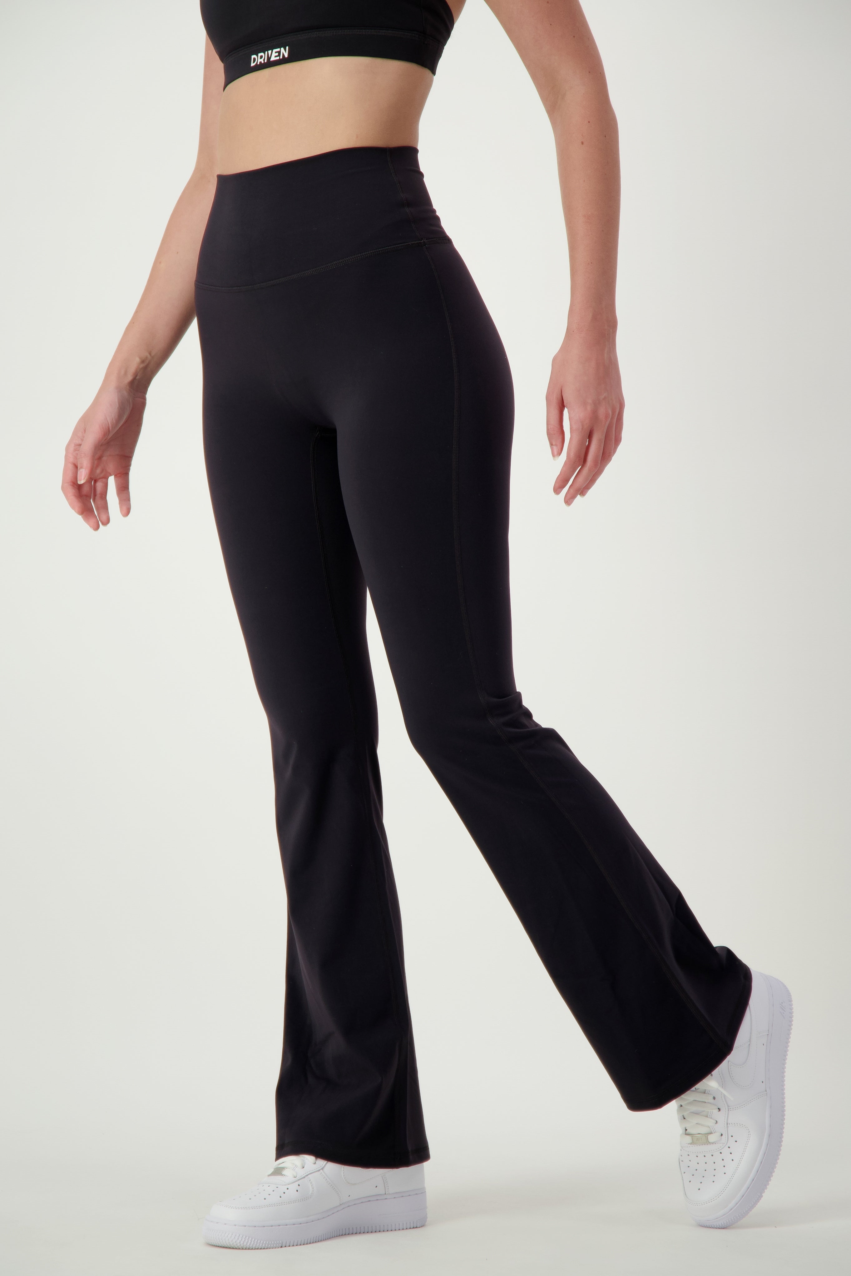 A person wearing black activewear, consisting of a sports bra and Classic Flare Tights (black) from DRIVEN Gym Apparel, along with white sneakers, stands against a plain white background. The person's head is not visible, and their left leg is slightly lifted, showcasing the fit and style of the fitness apparel.
