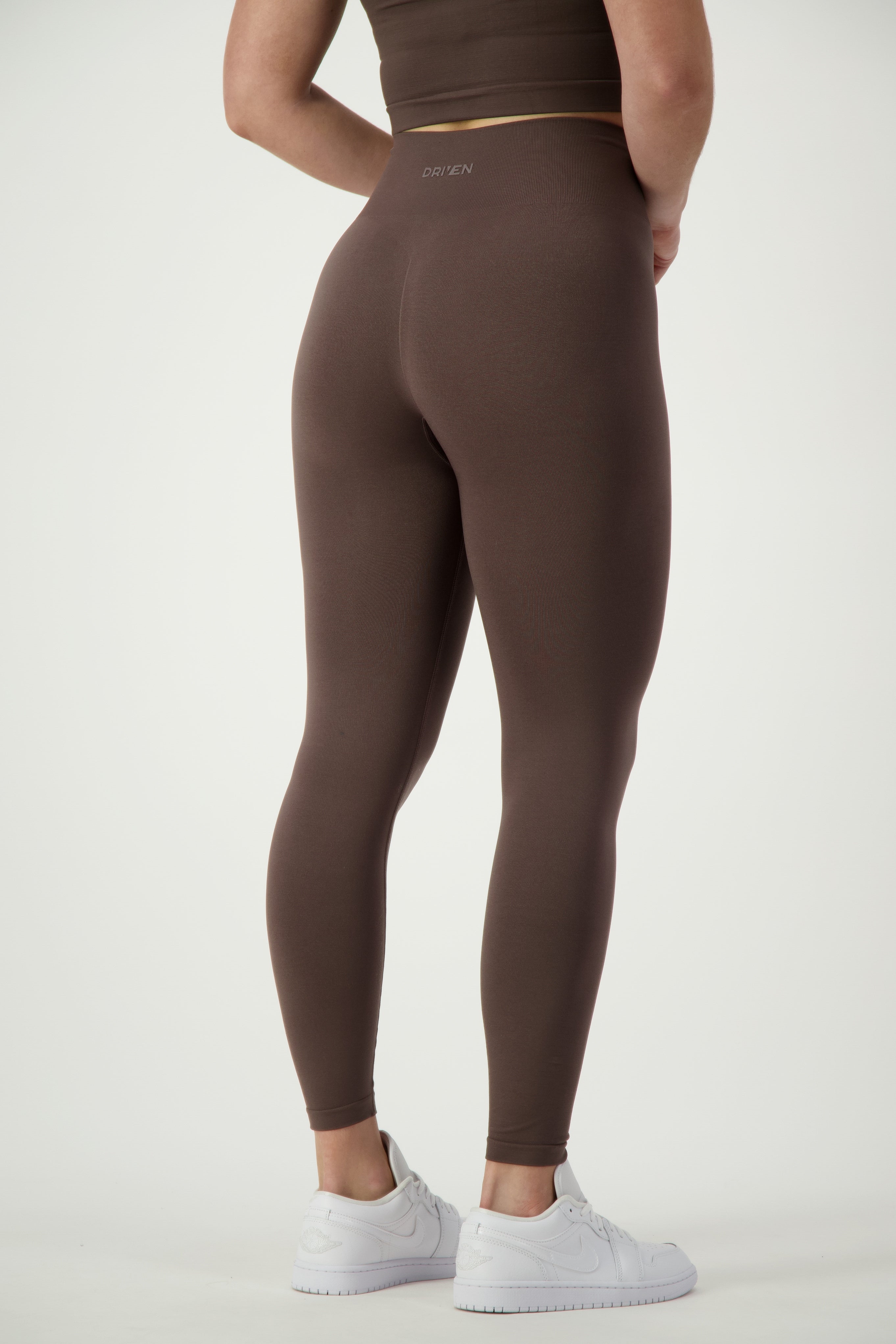 A person is shown from the back wearing a brown athletic outfit with "DRIVEN Gym Apparel" written on the waistband, epitomizing sleek fitness apparel. They are also sporting white sneakers. The background is plain white. The specific product they are wearing is the SL Flex Full Length Tights (mocha).