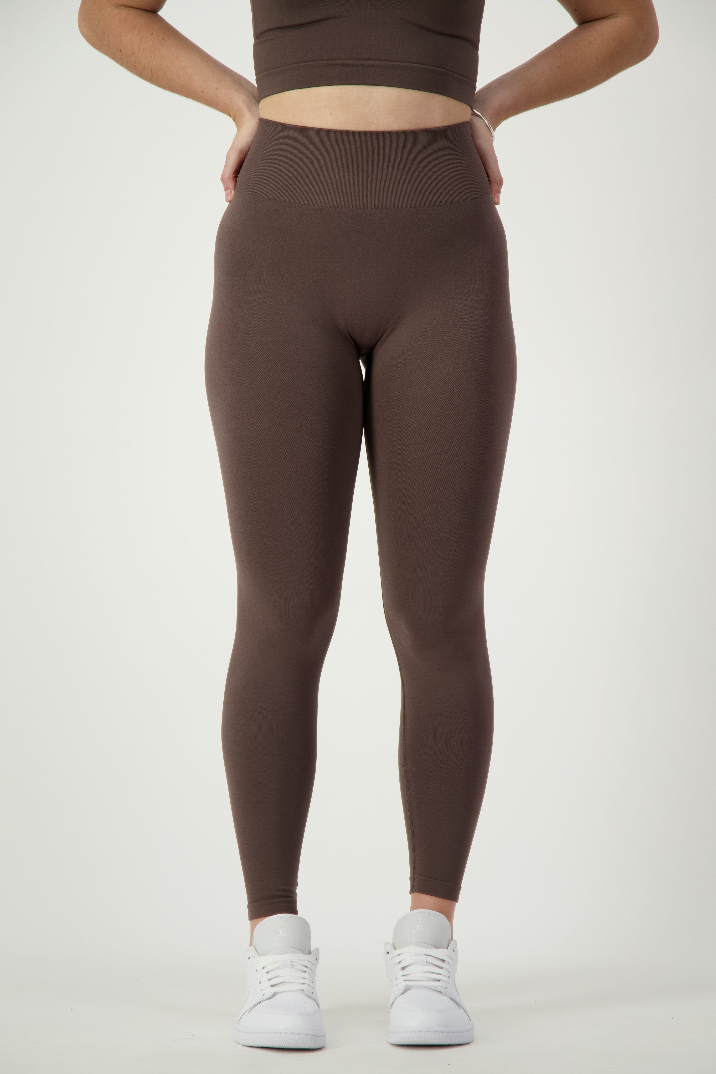 A person wearing a brown sports bra and matching SL Flex Full Length Tights (mocha) by DRIVEN Gym Apparel stands against a plain white background. The athleisure look is completed with white sneakers, hands placed confidently on their hips. Their head and upper torso are not shown.