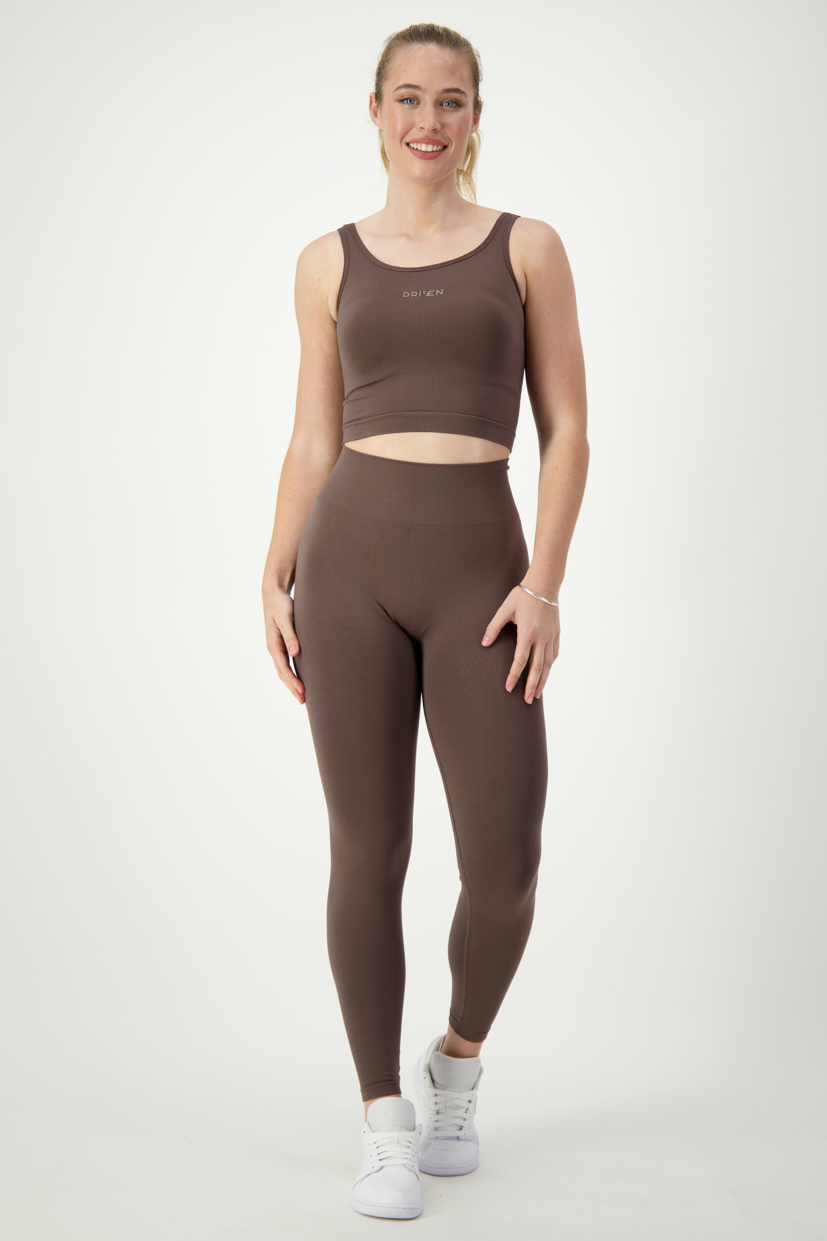 A person stands smiling against a plain background, showcasing trendy activewear. They are wearing a matching brown SL Flex Full Length Tights (mocha) from DRIVEN Gym Apparel, along with white sneakers. Their long hair is pulled back, and they appear relaxed and confident.