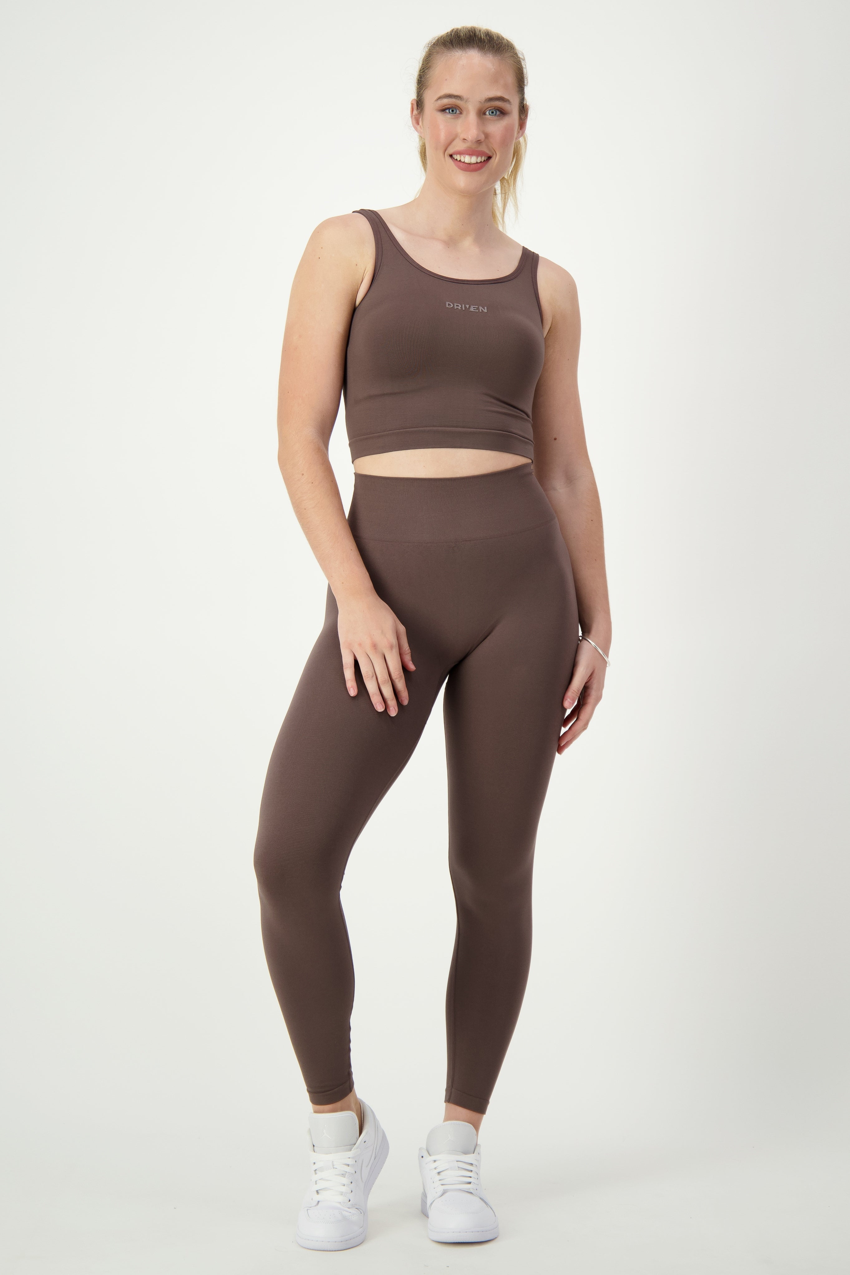 A person with long blonde hair is standing against a plain white background, wearing matching brown gym wear including a SL Flex Tank (mocha) by DRIVEN Gym Apparel and high-waisted leggings, paired with white sneakers. They are smiling and have one hand resting on their hip while the other hangs by their side.