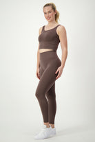A person stands confidently against a plain white background, wearing a matching brown fitness apparel set consisting of DRIVEN Gym Apparel's SL Flex Tank (mocha) and high-waisted leggings. They wear white sneakers and a bracelet, and they smile at the camera.