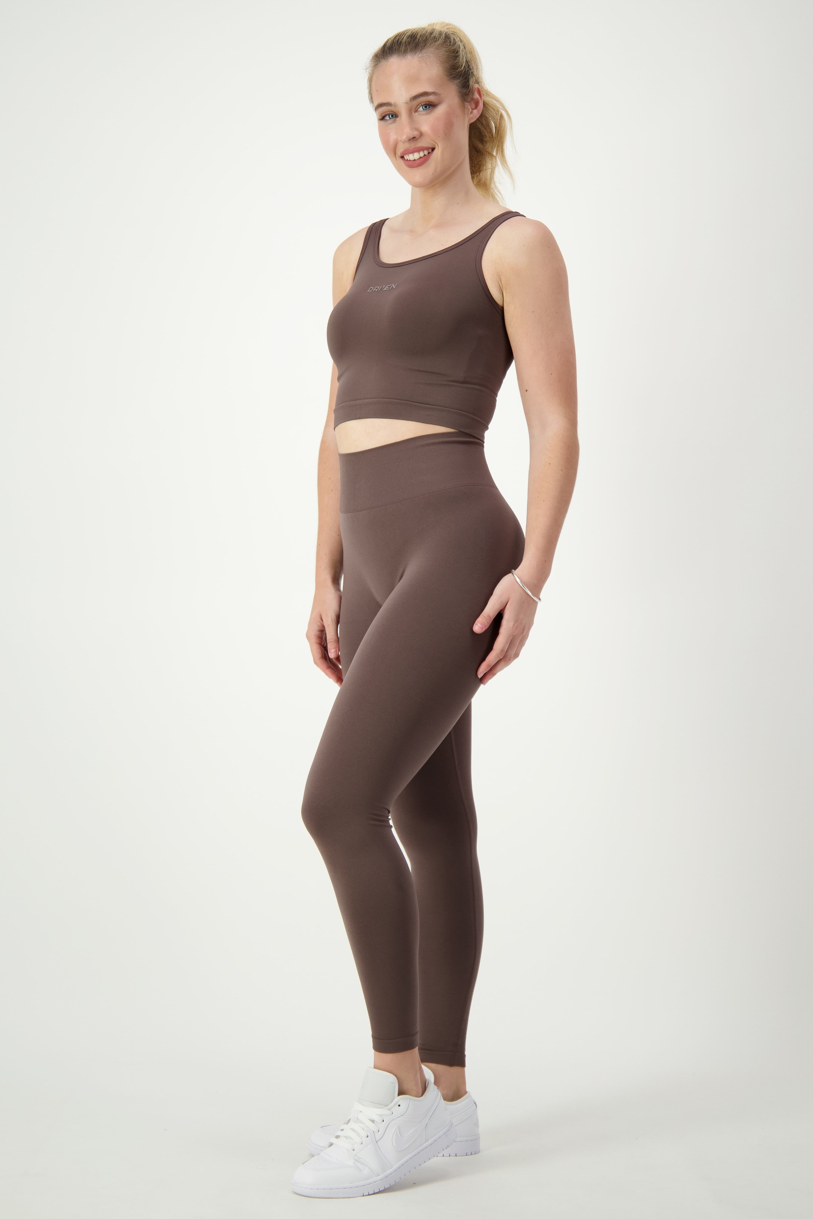 A person stands confidently against a plain white background, wearing a matching brown fitness apparel set consisting of DRIVEN Gym Apparel's SL Flex Tank (mocha) and high-waisted leggings. They wear white sneakers and a bracelet, and they smile at the camera.