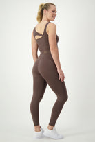 A woman stands in a light grey studio, wearing a brown sports bra and matching SL Flex Full Length Tights (mocha) by DRIVEN Gym Apparel, perfect athleisure. She is facing slightly to the side with her back partially to the camera. Her blonde hair is in a ponytail, and she is smiling while wearing white sneakers.