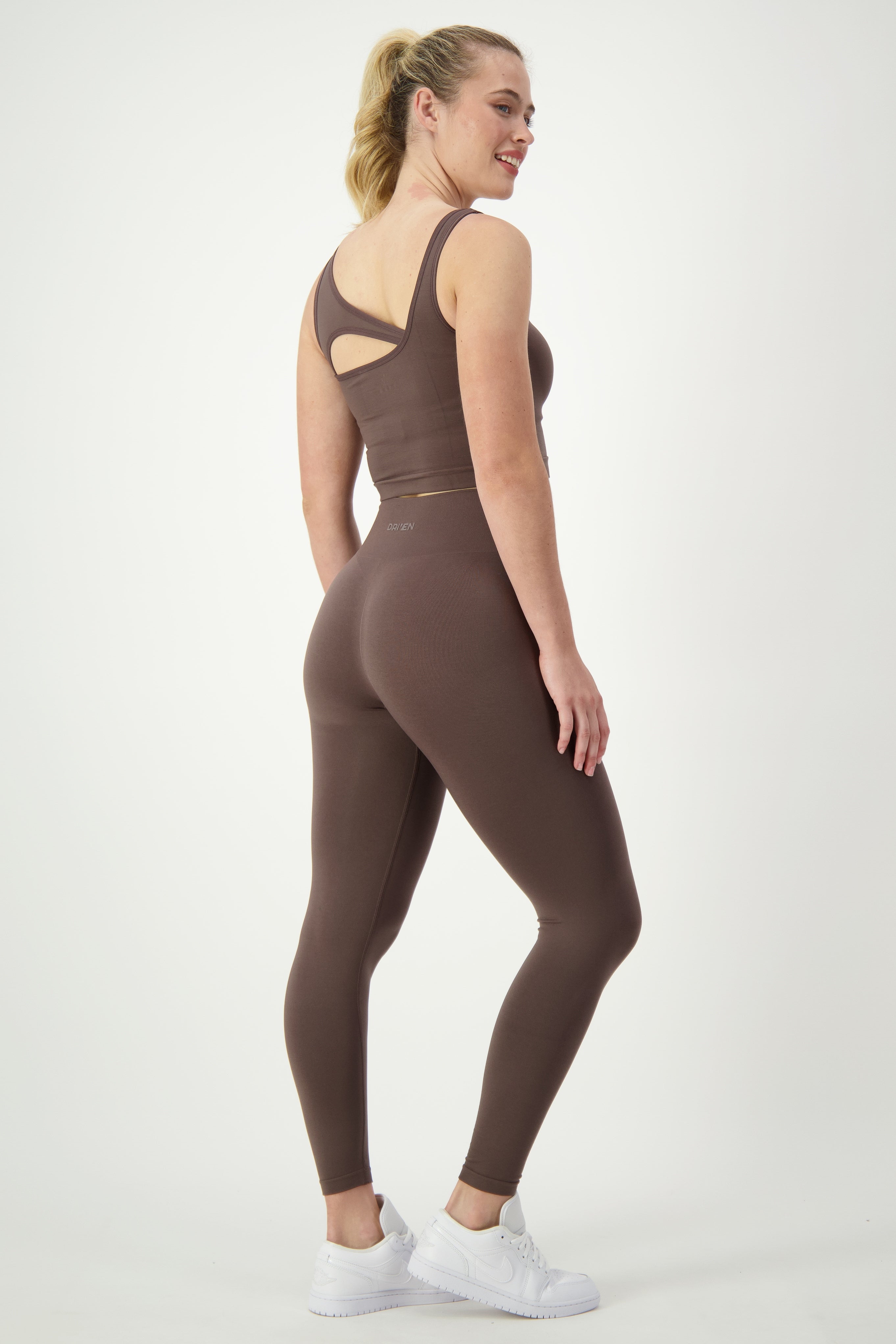 A woman stands in a light grey studio, wearing a brown sports bra and matching SL Flex Full Length Tights (mocha) by DRIVEN Gym Apparel, perfect athleisure. She is facing slightly to the side with her back partially to the camera. Her blonde hair is in a ponytail, and she is smiling while wearing white sneakers.
