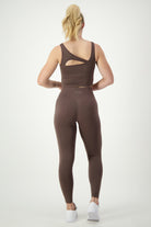 A person with a blonde ponytail is facing away, wearing a brown SL Flex Tank (mocha) and matching leggings as part of their athleisure outfit. The tank features a cutout detail on one shoulder, while "DRSKIN" is printed on the leggings' waistband. Completing the look are white athletic shoes.