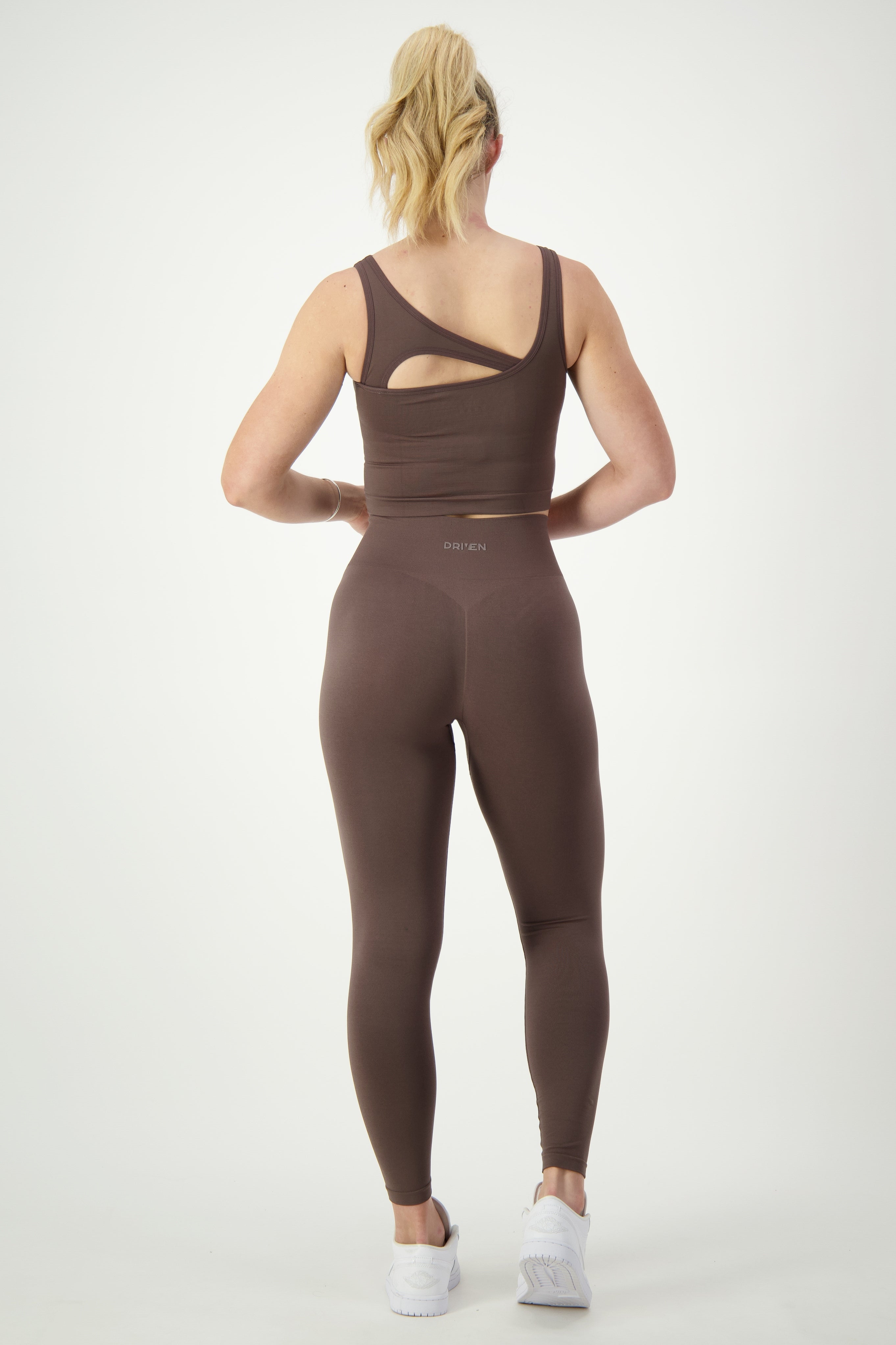 A person with a blonde ponytail is facing away, wearing a brown SL Flex Tank (mocha) and matching leggings as part of their athleisure outfit. The tank features a cutout detail on one shoulder, while "DRSKIN" is printed on the leggings' waistband. Completing the look are white athletic shoes.