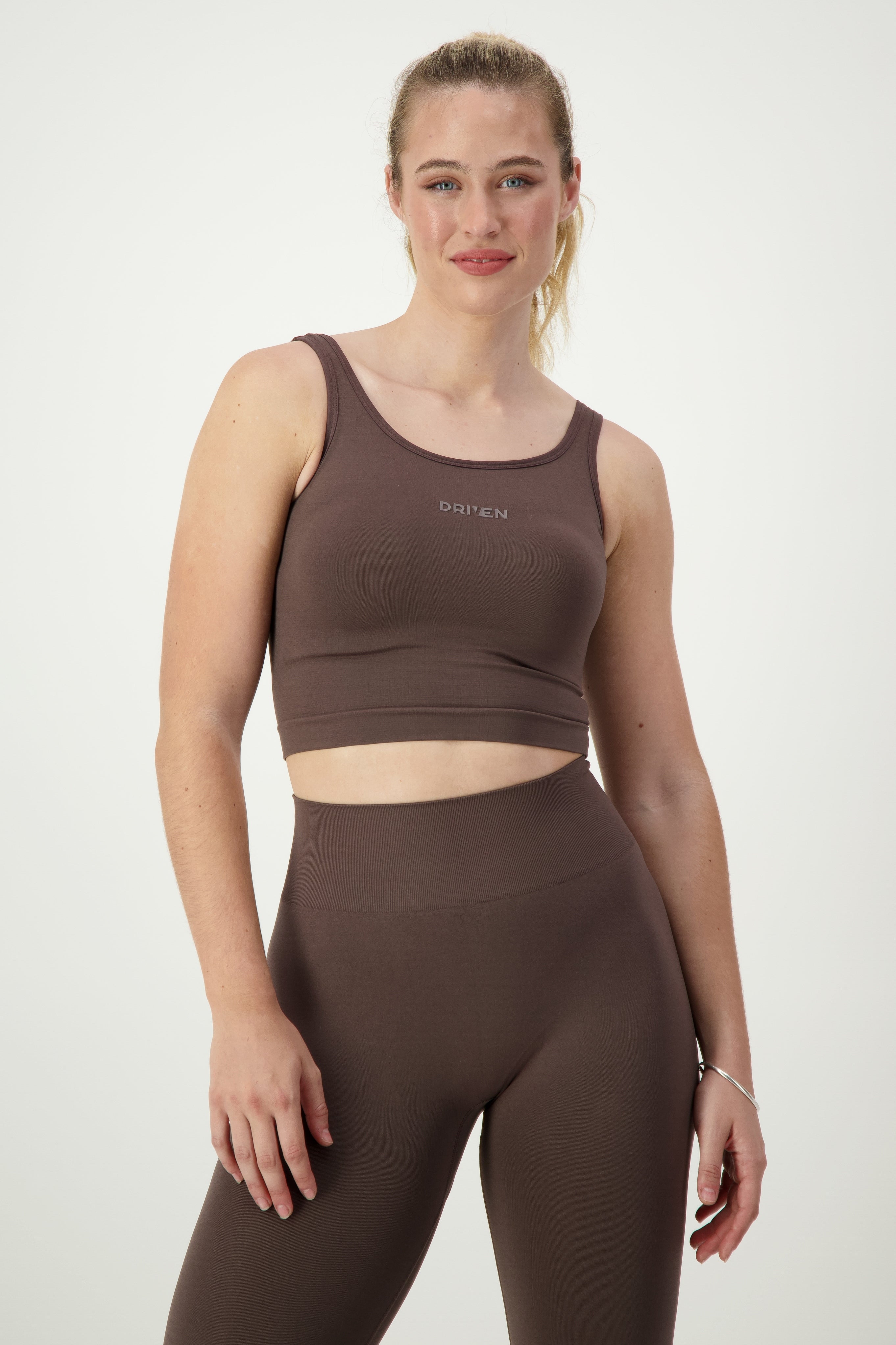 A person with long blond hair tied back is wearing a brown athletic crop top and matching high-waisted leggings, epitomizing stylish fitness apparel. They are standing against a plain white background and smiling slightly. The crop top has the word "DRIVEN" written on it, as it is the SL Flex Tank (mocha) from DRIVEN Gym Apparel.