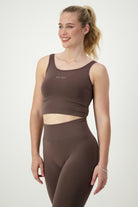 A woman with blonde hair in a high ponytail is wearing dark brown athleisure—a SL Flex Tank (mocha) by DRIVEN Gym Apparel and matching high-waisted leggings. She is standing and smiling, looking slightly to her left against a plain white background.