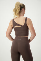 A woman with blonde hair is wearing a mocha SL Flex Tank and matching high-waisted leggings. She is standing with her back to the camera, showcasing the open-back design of the tank and the brand name "DRIVEN Gym Apparel" on the waistband of the activewear.