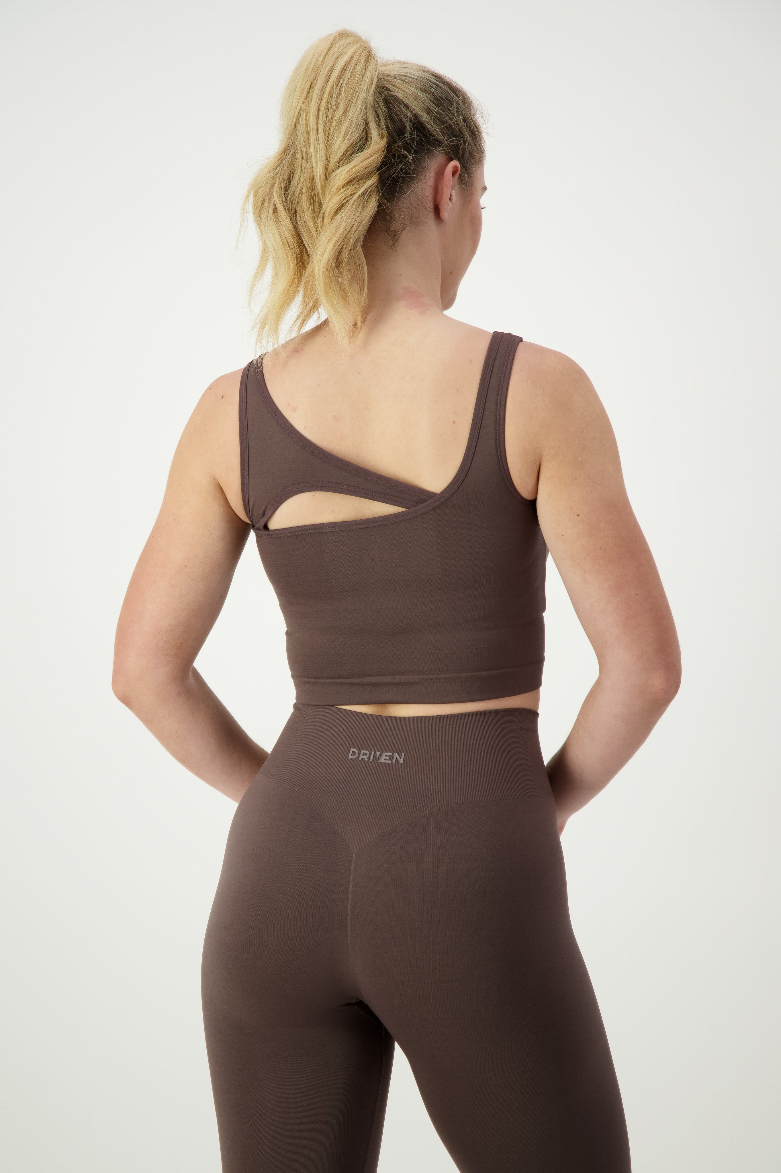A woman with blonde hair is wearing a mocha SL Flex Tank and matching high-waisted leggings. She is standing with her back to the camera, showcasing the open-back design of the tank and the brand name "DRIVEN Gym Apparel" on the waistband of the activewear.