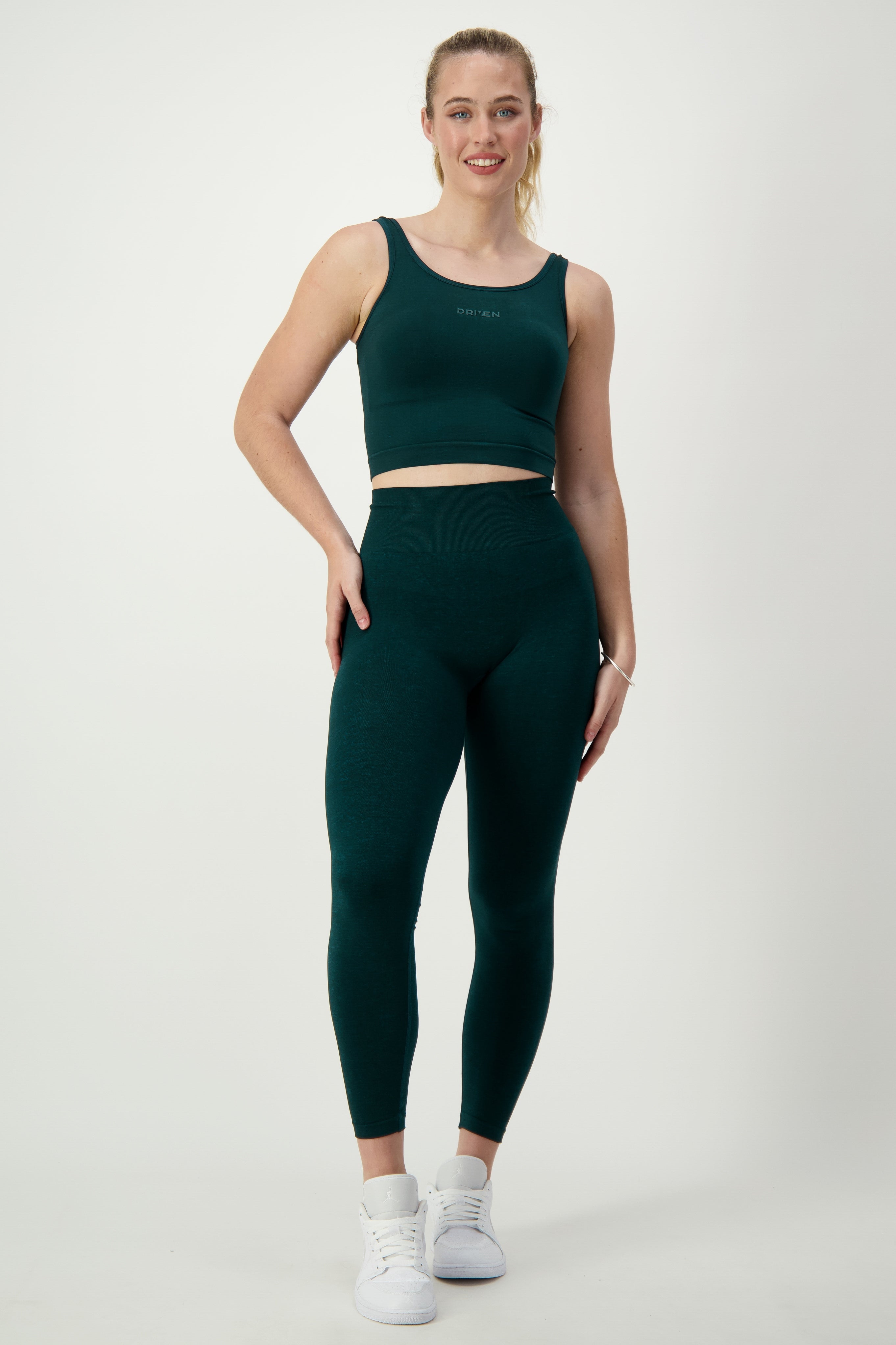 A woman is standing against a white background, wearing dark green activewear that includes a sleeveless crop top and high-waisted DRIVEN Gym Apparel SL Flex Full Length Tights (sea green). She has her hair tied back and is smiling while sporting white athletic shoes.