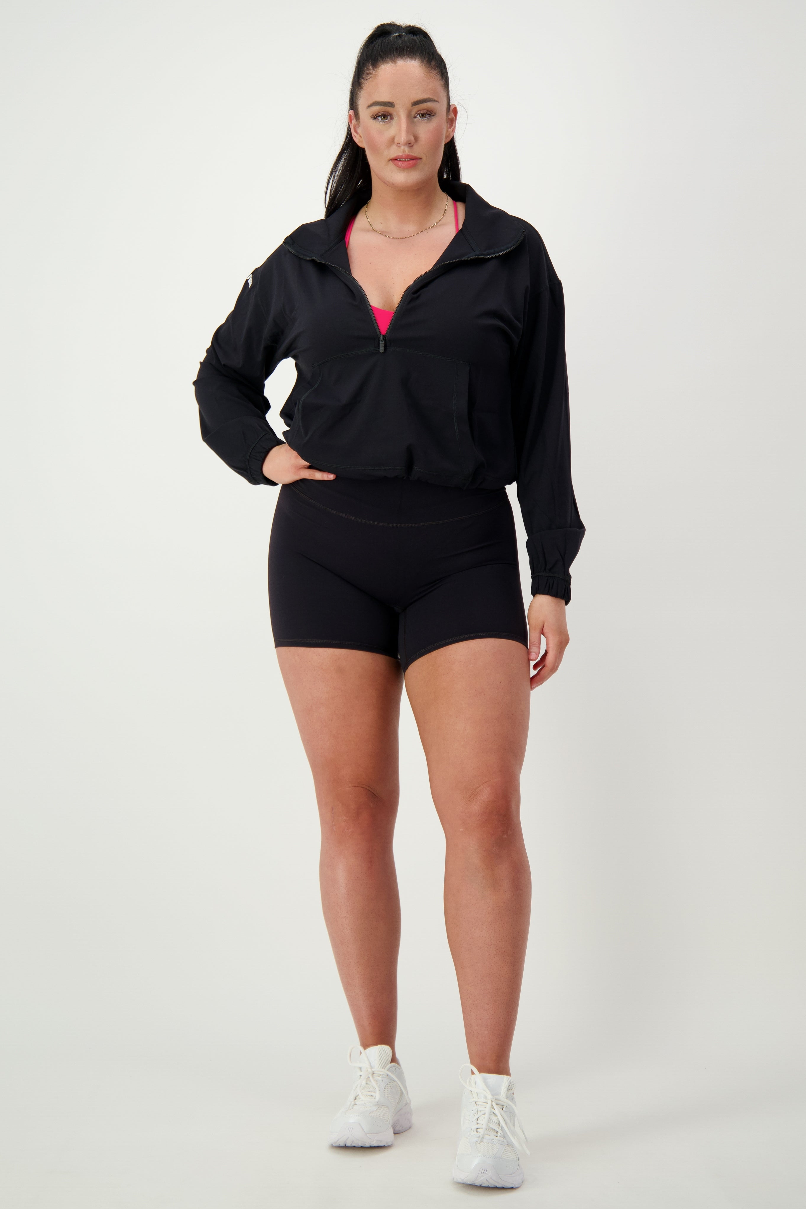 A woman stands against a plain background wearing black activewear: a long-sleeve jacket, black shorts, and white athletic shoes. The Lara Half Zip Jacket (black) from DRIVEN Gym Apparel is unzipped slightly to reveal a glimpse of a pink sports bra underneath. She has her hair tied back in a ponytail and faces the camera directly.