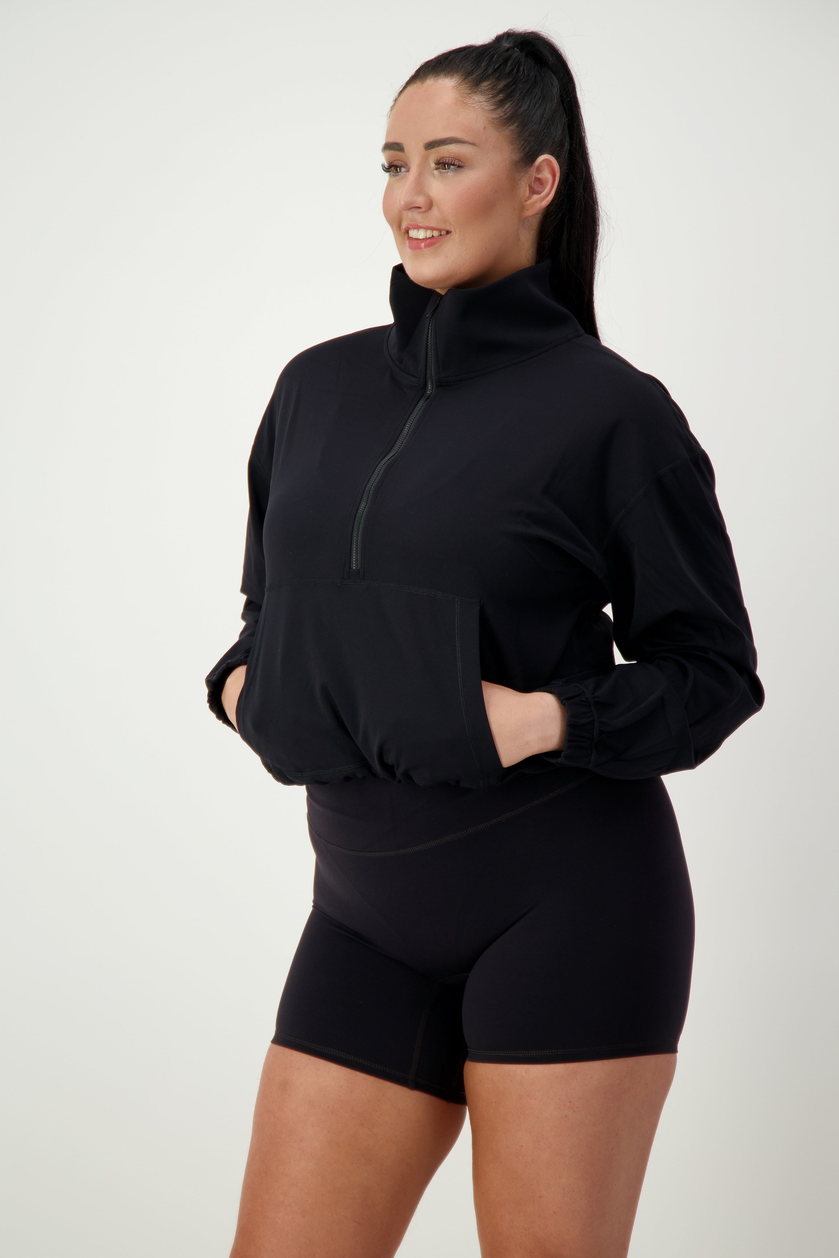 A person with long dark hair is wearing black athleisure, consisting of a DRIVEN Gym Apparel Lara Half Zip Jacket (black) and black shorts. They are standing with hands in the jacket's front pockets, smiling and looking to the right against a plain white background.