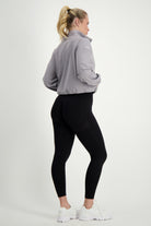 A person with blonde hair in a ponytail is wearing the Lara Half Zip Jacket (light grey) by DRIVEN Gym Apparel, black leggings, and white sneakers. The person is standing with their back turned, glancing over one shoulder against a plain white background.