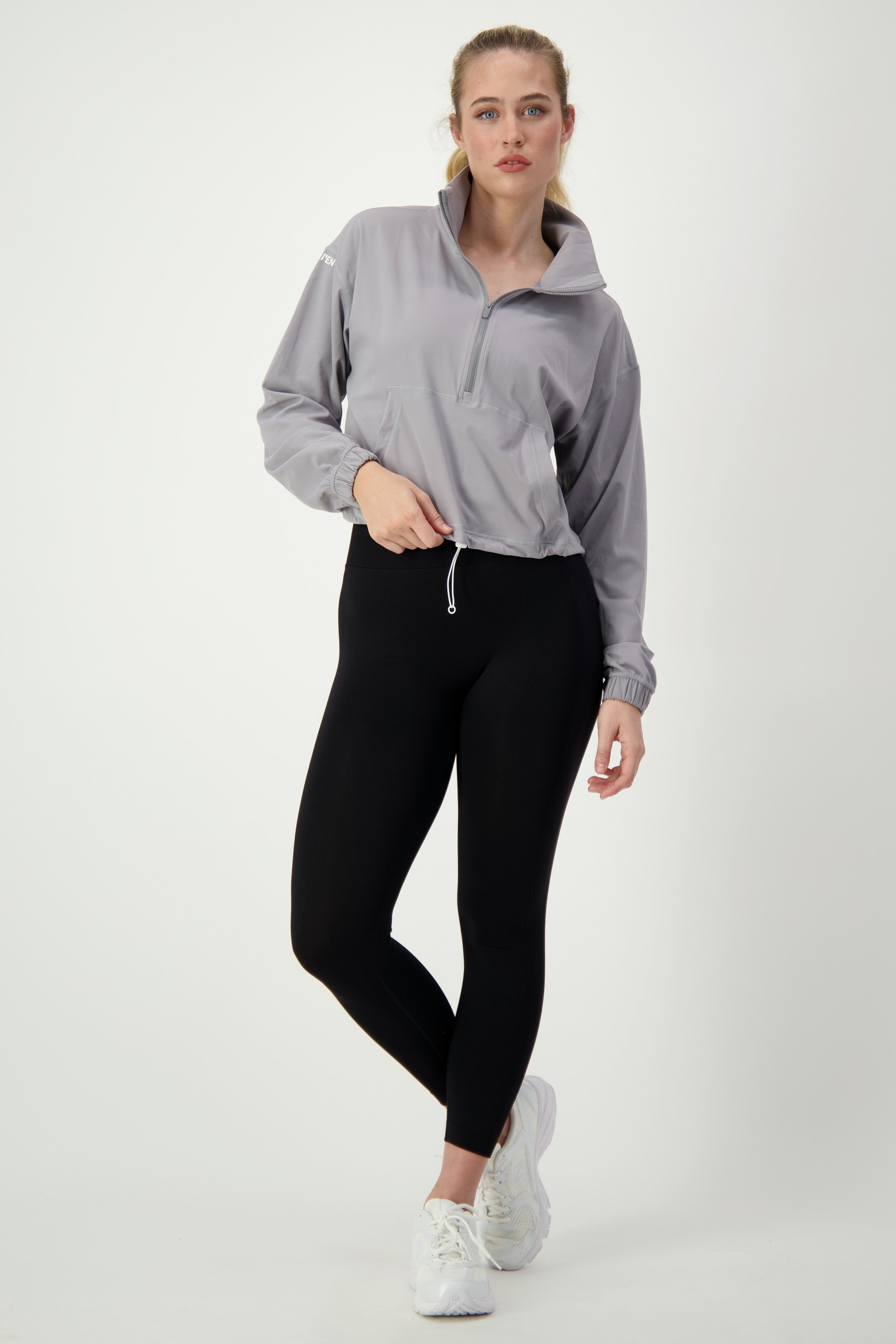 A person with light hair tied back, wearing a light gray Lara Half Zip Jacket from DRIVEN Gym Apparel, black leggings, and white sneakers, is standing against a plain white background. They embody casual Athleisure style by slightly leaning to one side with one hand in their pocket.