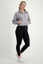A woman stands against a plain white background. She is wearing light gray fitness apparel, including the Lara Half Zip Jacket (light grey) by DRIVEN Gym Apparel with the collar up and black leggings. Her white athletic shoes complement her gym wear, and her blonde hair is tied back in a ponytail. She looks directly at the camera with a neutral expression.