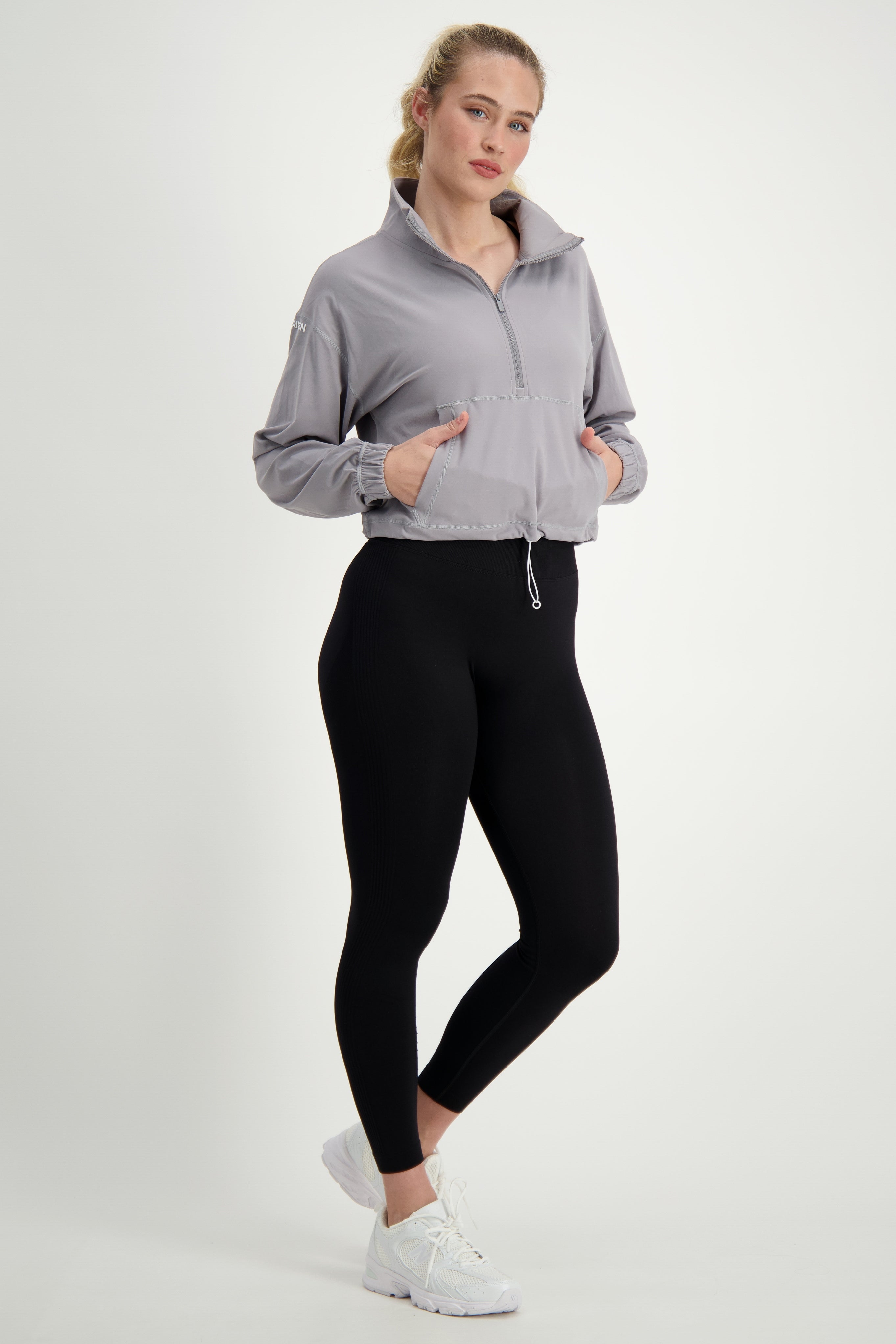 A woman stands against a plain white background. She is wearing light gray fitness apparel, including the Lara Half Zip Jacket (light grey) by DRIVEN Gym Apparel with the collar up and black leggings. Her white athletic shoes complement her gym wear, and her blonde hair is tied back in a ponytail. She looks directly at the camera with a neutral expression.