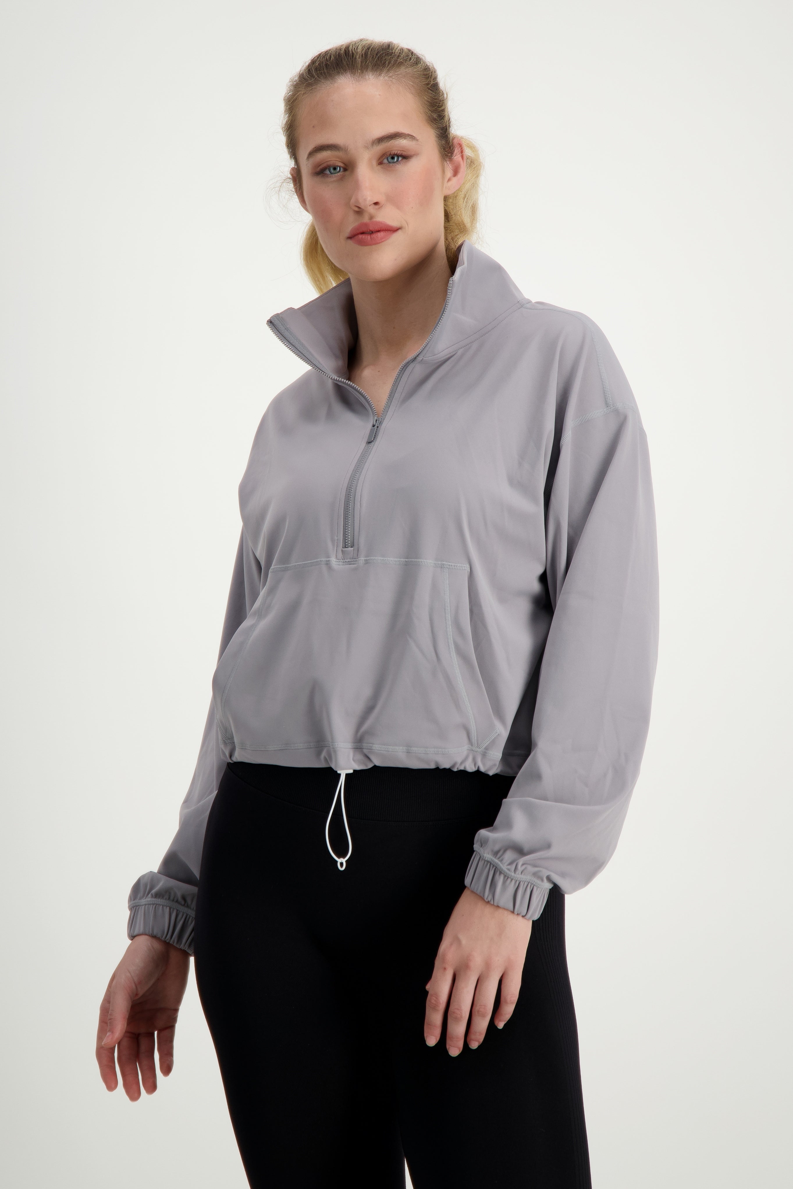 A person stands against a plain white background wearing a light gray Lara Half Zip Jacket (light grey) by DRIVEN Gym Apparel with a half-zip front and drawstring hem, embodying athleisure. Paired with black leggings, this activewear look is completed by their blond hair pulled back as they pose with a neutral expression.