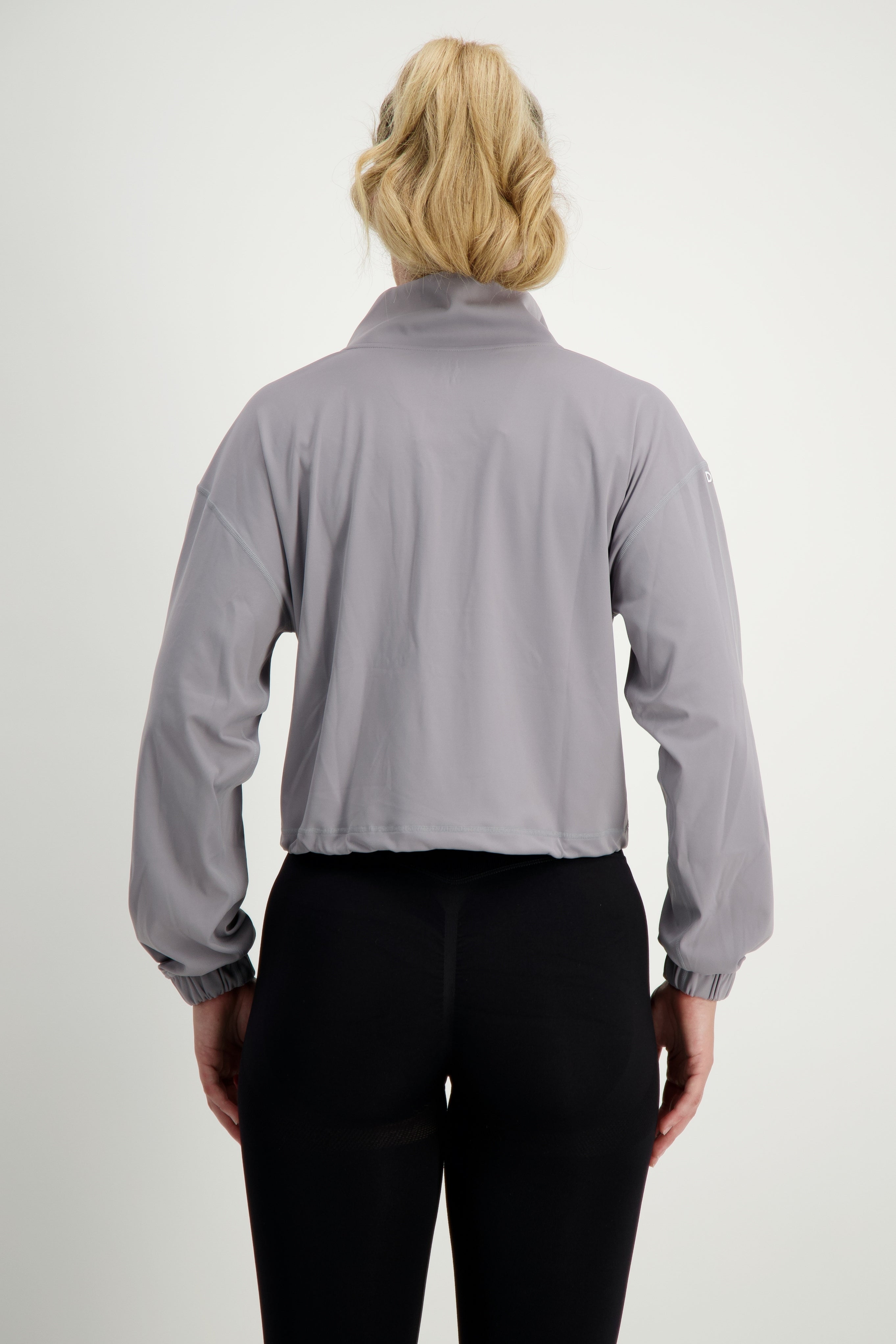 A person with blonde hair tied in a ponytail is shown from the back, wearing a Lara Half Zip Jacket (light grey) by DRIVEN Gym Apparel and black leggings, epitomizing stylish gym wear. The background is plain white.