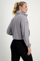 A person with long blonde hair in a ponytail is standing sideways while facing left. They are wearing a light grey Lara Half Zip Jacket from DRIVEN Gym Apparel with a high collar and black leggings, epitomizing athleisure style. The jacket has the text "DRIVEN" on the upper left sleeve against a plain white background.