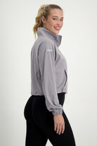 A woman with her hair in a ponytail smiles at the camera while standing sideways. She is wearing a light gray Lara Half Zip Jacket (light grey) by DRIVEN Gym Apparel and black leggings. The plain white background emphasizes her casual athleisure attire, perfect for gym wear or running errands.
