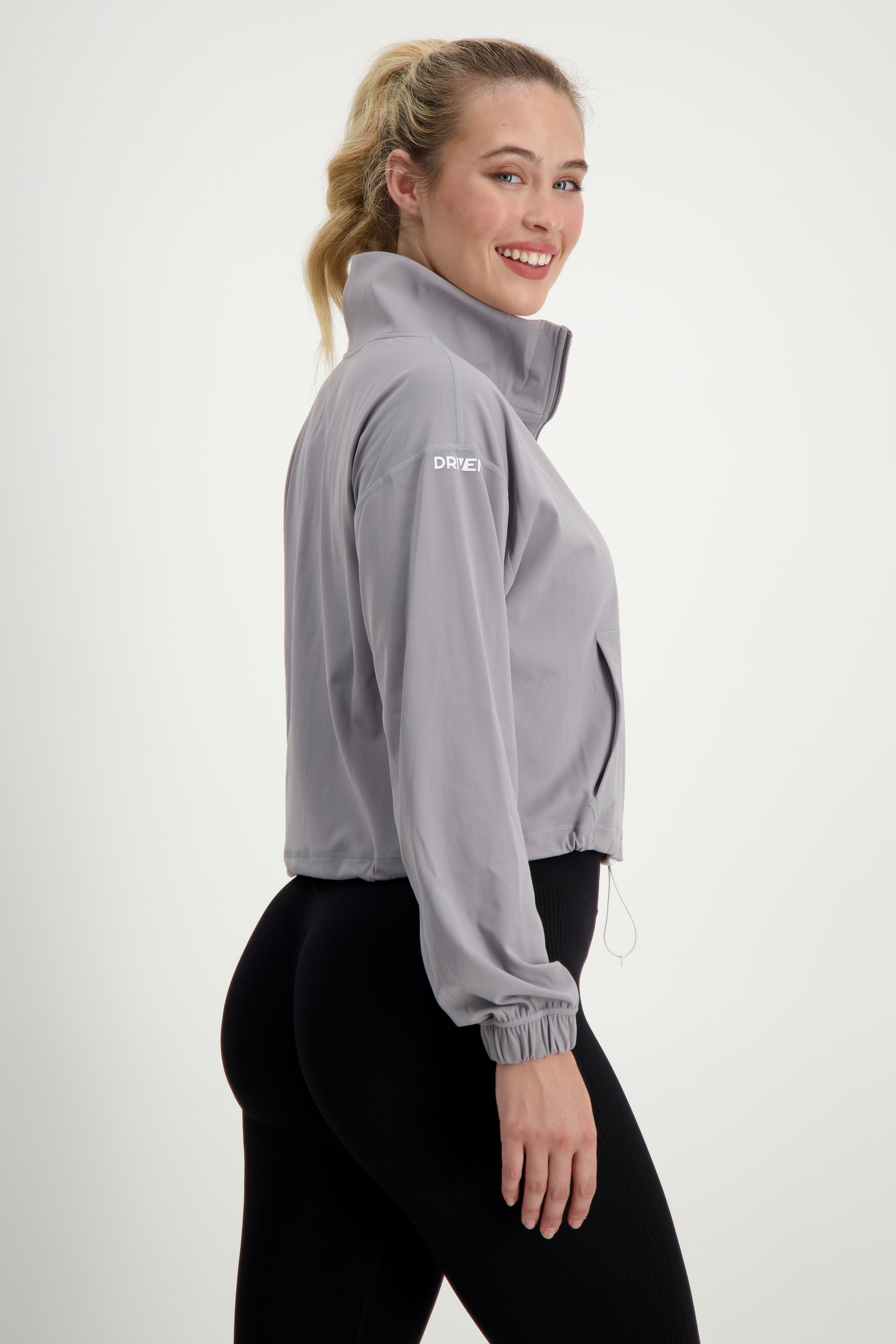 A woman with her hair in a ponytail smiles at the camera while standing sideways. She is wearing a light gray Lara Half Zip Jacket (light grey) by DRIVEN Gym Apparel and black leggings. The plain white background emphasizes her casual athleisure attire, perfect for gym wear or running errands.