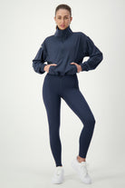 A person stands against a plain white background, wearing a dark blue Lara Half Zip Jacket (navy) by DRIVEN Gym Apparel and matching leggings that epitomize athleisure. They are also sporting white sneakers. The individual has one hand on their hip and a neutral expression.